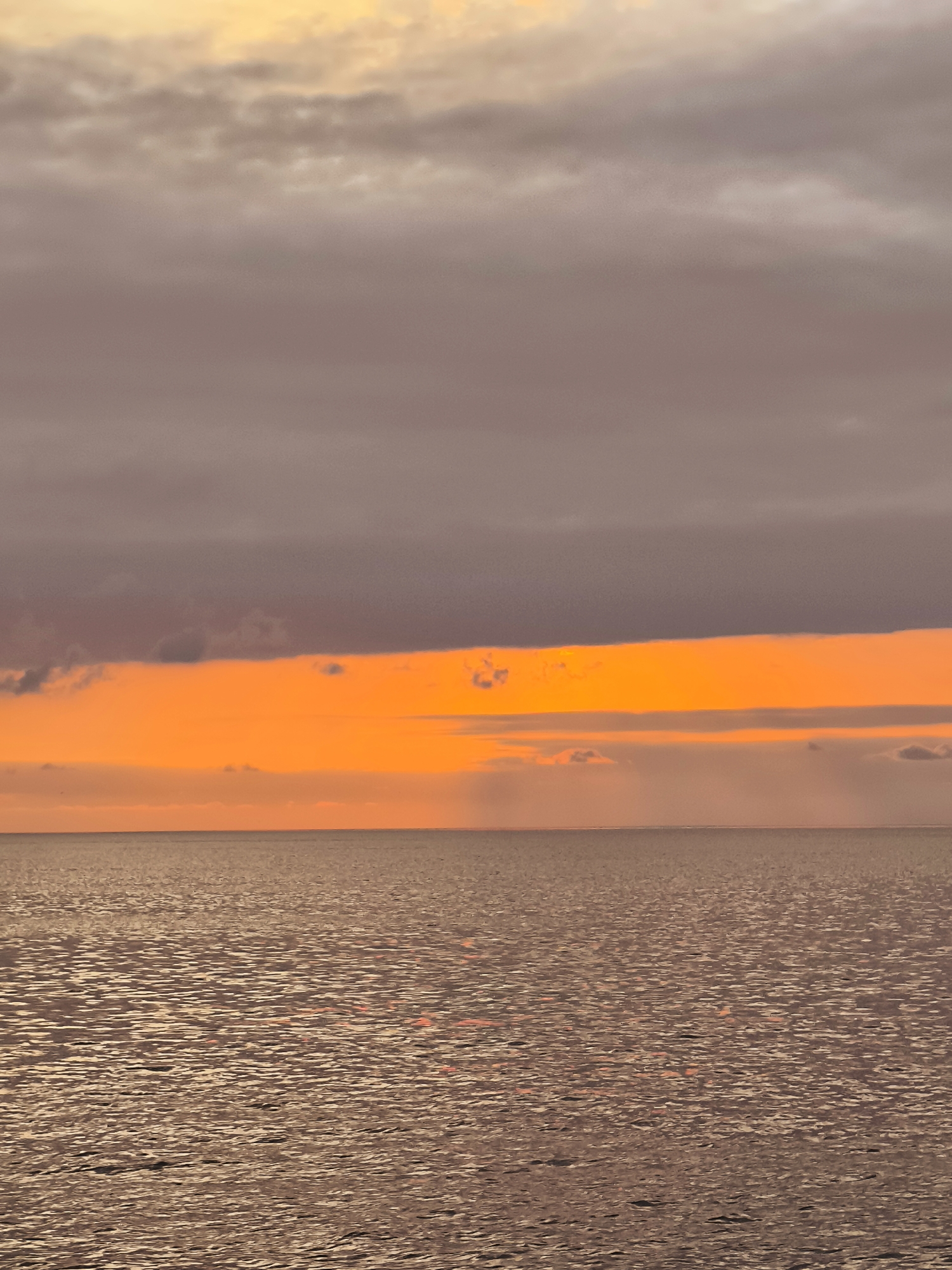 The image depicts a serene seascape at dusk, likely along the coast of Batumi, Georgia. The scene is dominated by the horizon line separating a calm sea from a dramatically colored sky. The sea, occupying the lower half of the frame, is a textured expanse of muted, shimmering browns and grays, reflecting the soft light of the setting sun. The water's surface is rippled, creating a mosaic of light and shadow.
Above the sea, a vibrant band of intense orange light cuts across the horizon, suggesting the sun is just below the visible edge. This bright orange layer is overlain by softer, diffused shades of pink and pale yellow, indicative of scattered clouds catching the last rays of daylight. Further up, the sky transitions into darker, muted grays and purples, with scattered, indistinct clouds. There are no people, boats, or landmasses visible in the image, creating a sense of vastness and tranquility. The overall atmosphere is peaceful and contemplative, capturing the fleeting beauty of a coastal sunset.