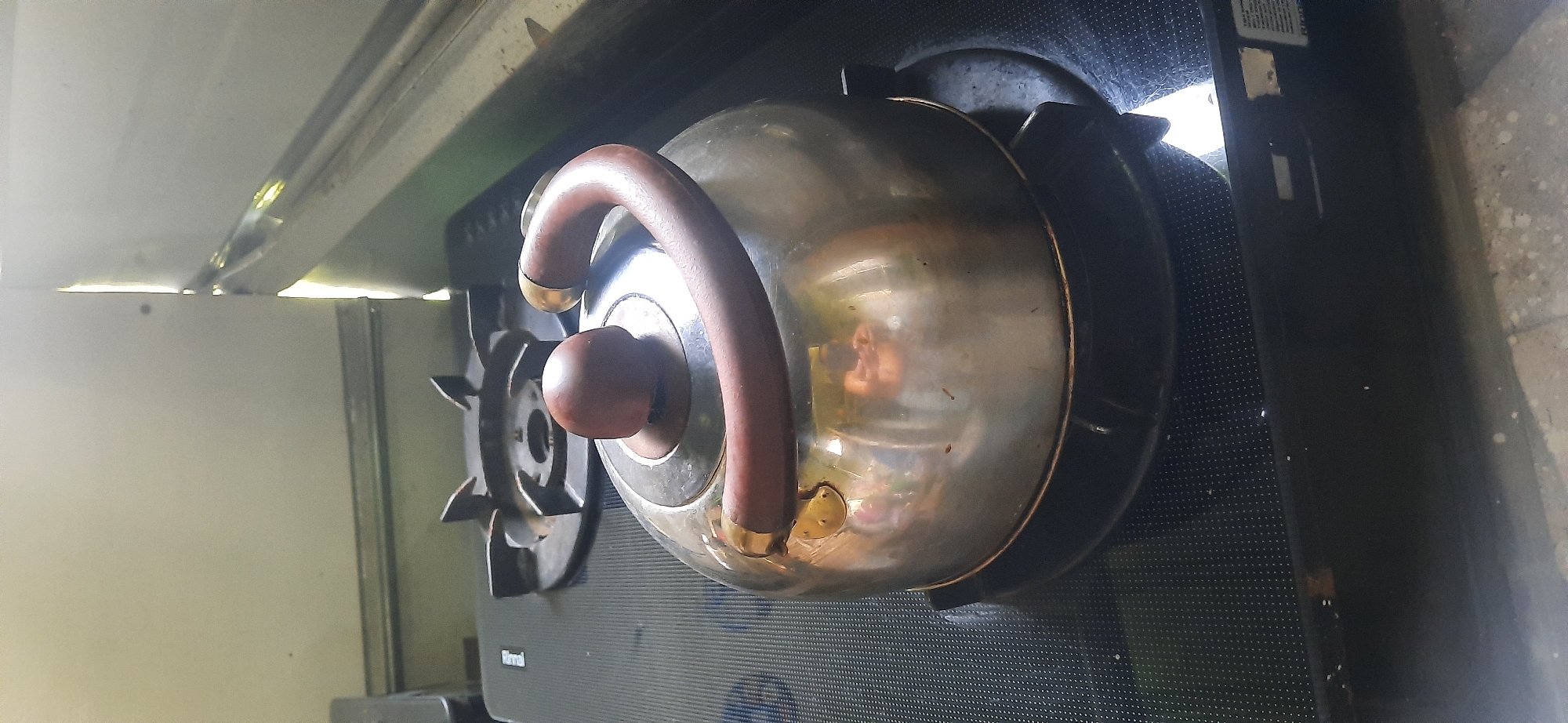 A shiny metal kettle with a dark brown handle and lid sits on a black gas stovetop. The kettle is round with a slightly flared bottom. The stovetop has a textured surface with small circular indentations. A gas burner grate is visible behind the kettle, appearing to be made of dark metal. The background is a light-colored wall with a bright light source shining from the top right. The scene appears to be in a kitchen.