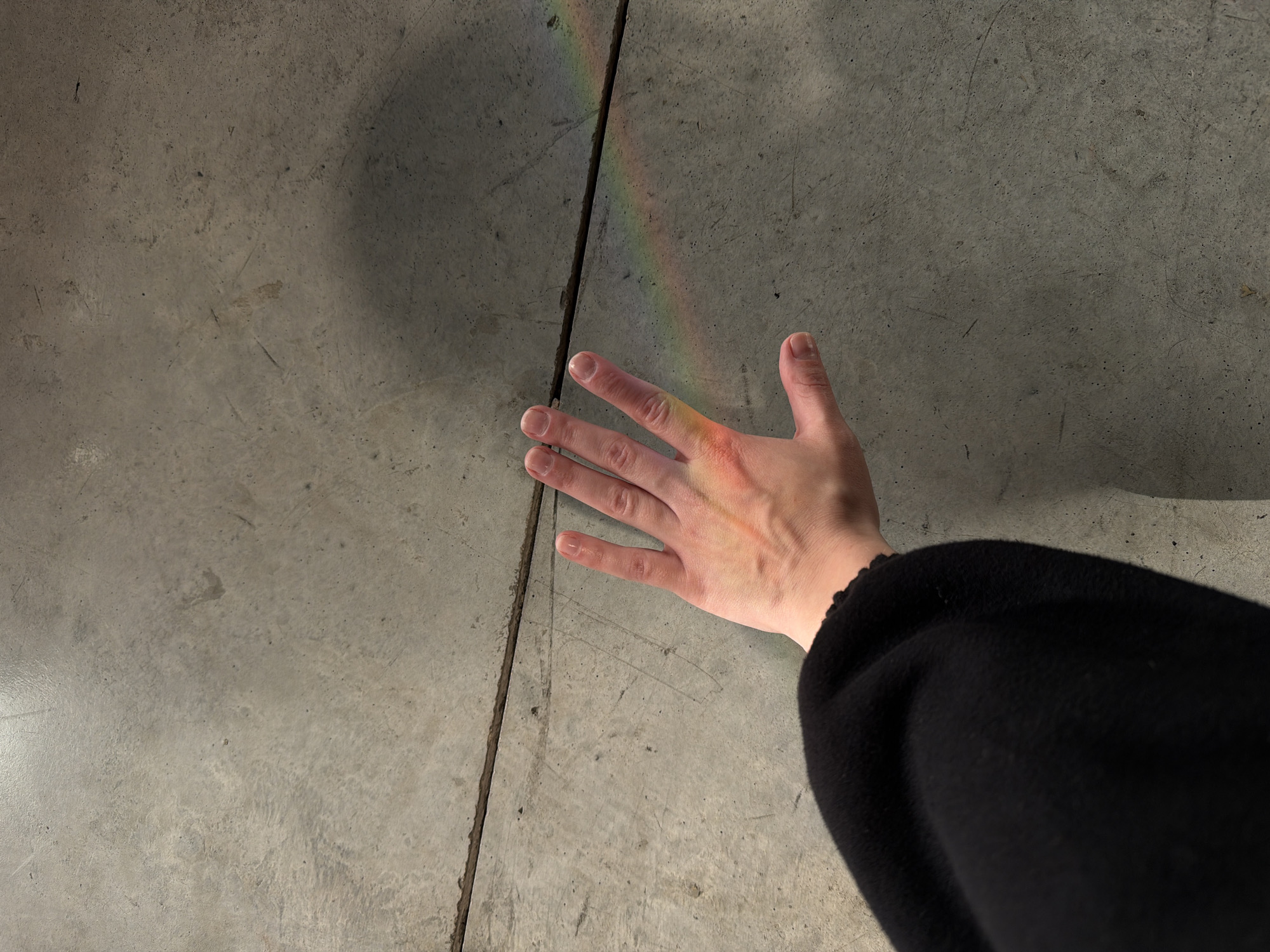 A top-down shot captures a person's hand resting on a gray concrete surface, possibly a floor. A narrow band of refracted light, resembling a rainbow, runs diagonally across the concrete and falls across the hand. The person wears a black garment, only a portion of which is visible in the lower right corner of the image. There are no other discernable people, objects, actions, or interactions. The location in Tel-Aviv, Israel cannot be determined from the image.