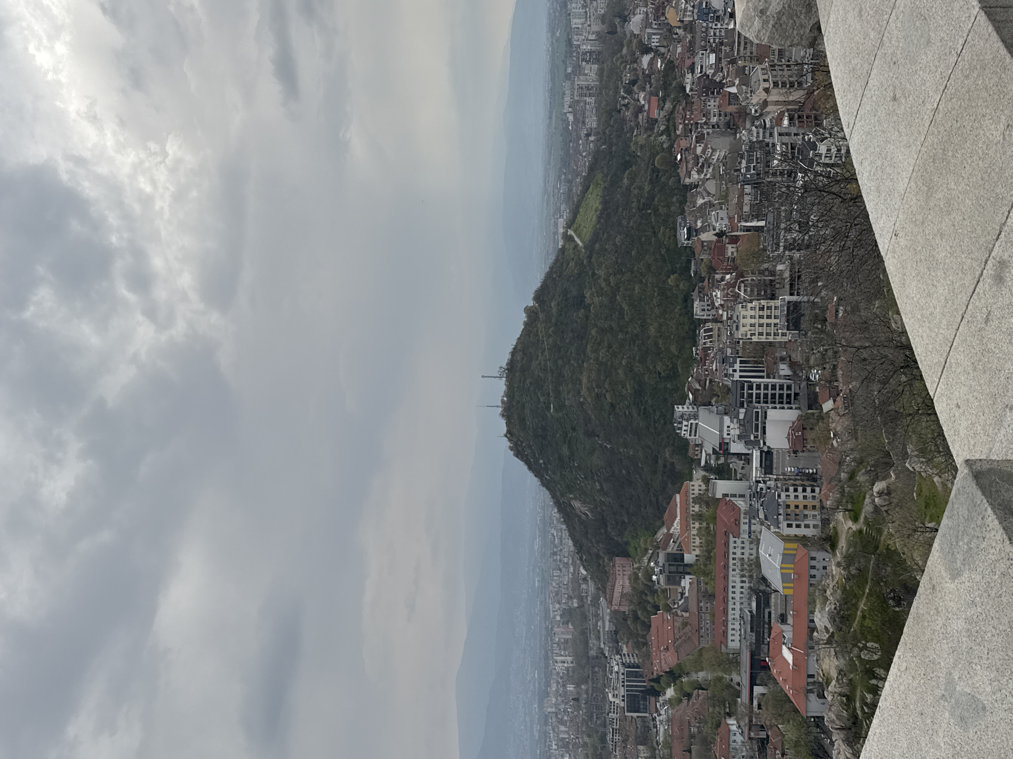 The image shows a cloudy, overcast day overlooking the city of Plovdiv, Bulgaria. A large, tree-covered hill dominates the center of the frame, with the city sprawling below and beyond it. In the distance, hazy mountains fade into the horizon. A gray stone structure is visible in the right foreground. Radio masts are seen atop the hill. Buildings of various sizes and architectural styles, with predominantly orange and red roofs, densely pack the urban landscape. The sky is gray with heavy clouds.