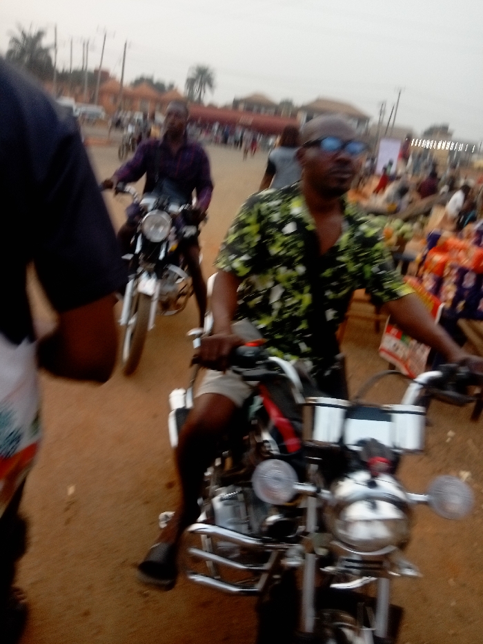 The image depicts an outdoor street or market scene, likely in a town or village, characterized by a reddish-brown unpaved ground. The overall impression is one of daily activity and movement.
In the immediate foreground, to the left, the shoulder, arm, and part of the torso of a person wearing a dark blue or black short-sleeved top and white or patterned shorts are visible.
The central and most in-focus subject is a man riding a motorcycle. He is wearing a short-sleeved, collared shirt with a black base and a prominent, abstract pattern of light green or yellowish shapes. He is also wearing light-colored shorts and dark slippers or sandals. He has short hair, a short beard or stubble, and is wearing dark-rimmed glasses with a distinct blue tint on the lenses, reflecting some light. His hands are on the chrome handlebars of his motorcycle, which features a large round headlight, side mirrors, and what appears to be a chrome crash bar or engine guard at the front.
Behind this man and slightly to his left, another man is also on a motorcycle. He is wearing a dark purple long-sleeved shirt and dark shorts. A dark bag is slung across his chest. His face and the details of his motorcycle are blurrier than the foreground subject, suggesting either motion or a shallower depth of field. This motorcycle also has a noticeable round headlight and chrome accents.
The background is significantly blurred, indicating motion or a distant focus. Visible elements in the background include low-rise buildings with reddish or terracotta-colored roofs, suggestive of local architecture. Power lines and utility poles are faintly discernible against the hazy sky. To the far right in the background, there are hints of colorful items, possibly textiles or produce, arranged on what might be market stalls, with shades of orange and red being prominent. A few palm trees are also visible in the distance.
The lighting suggests it is daytime, possibly late afternoon or an overcast day, as there are no harsh shadows. The general blurriness, especially of the background, indicates either camera shake, motion blur of the subjects, or a quick snapshot. No readable text is visible in the image.