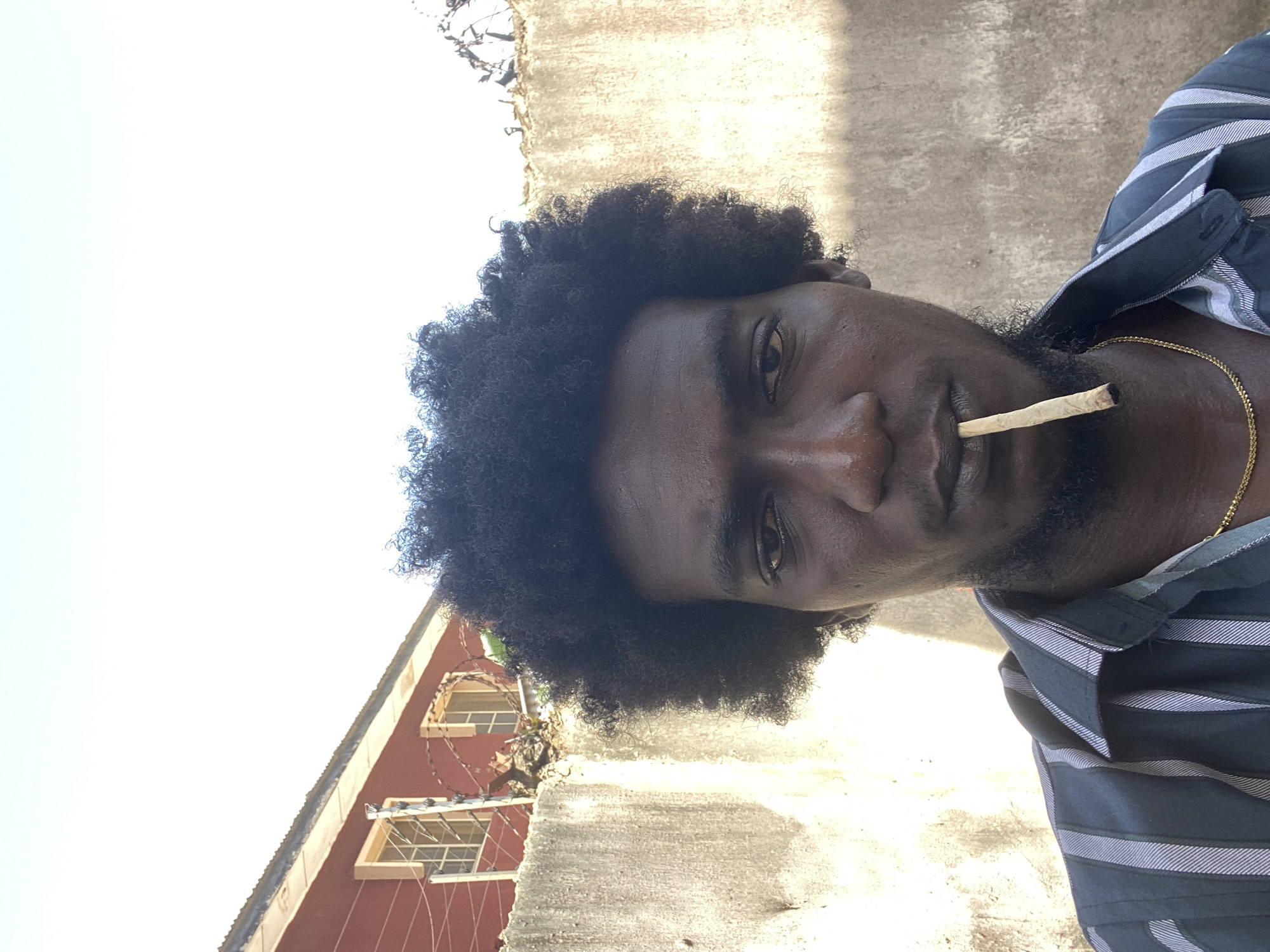 A close-up, rotated, angled shot of a dark-skinned man with an Afro hairstyle and a thin beard, holding what appears to be a rolled cigarette in his mouth. He is wearing a dark, striped shirt and a gold chain. The background features a weathered concrete wall and a building with visible windows, possibly in Lagos, Nigeria. The overall lighting suggests a bright, sunny day.