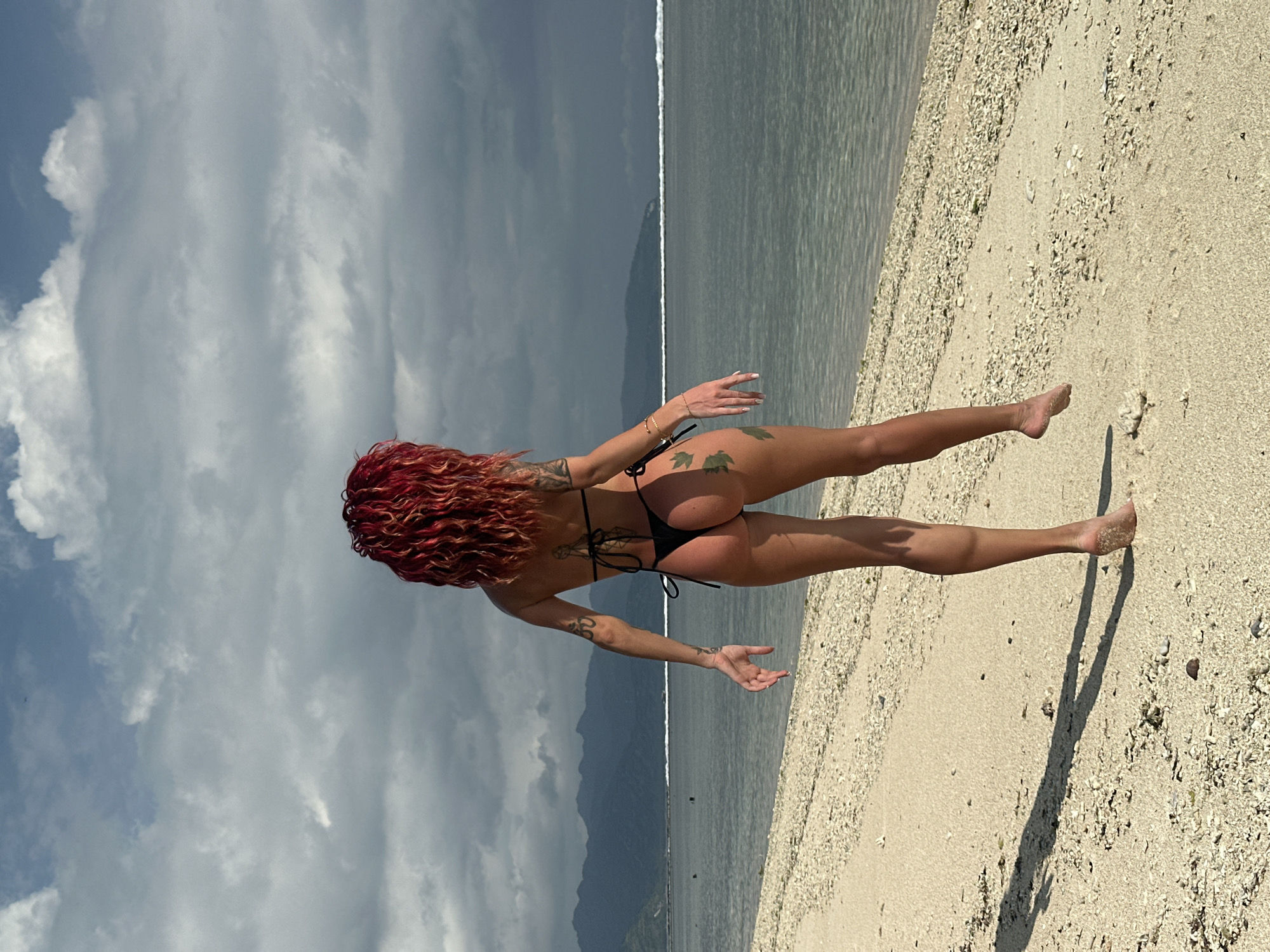 The image shows a woman with red curly hair, facing away from the camera, wearing a black bikini and standing on a beach. The woman has tattoos on her arms and back. The beach has light-colored sand or small shells. Behind the beach is the ocean, and in the background is a mountain range under a cloudy sky. The location appears to be a beach near mountains, in Unknown, Indonesia.