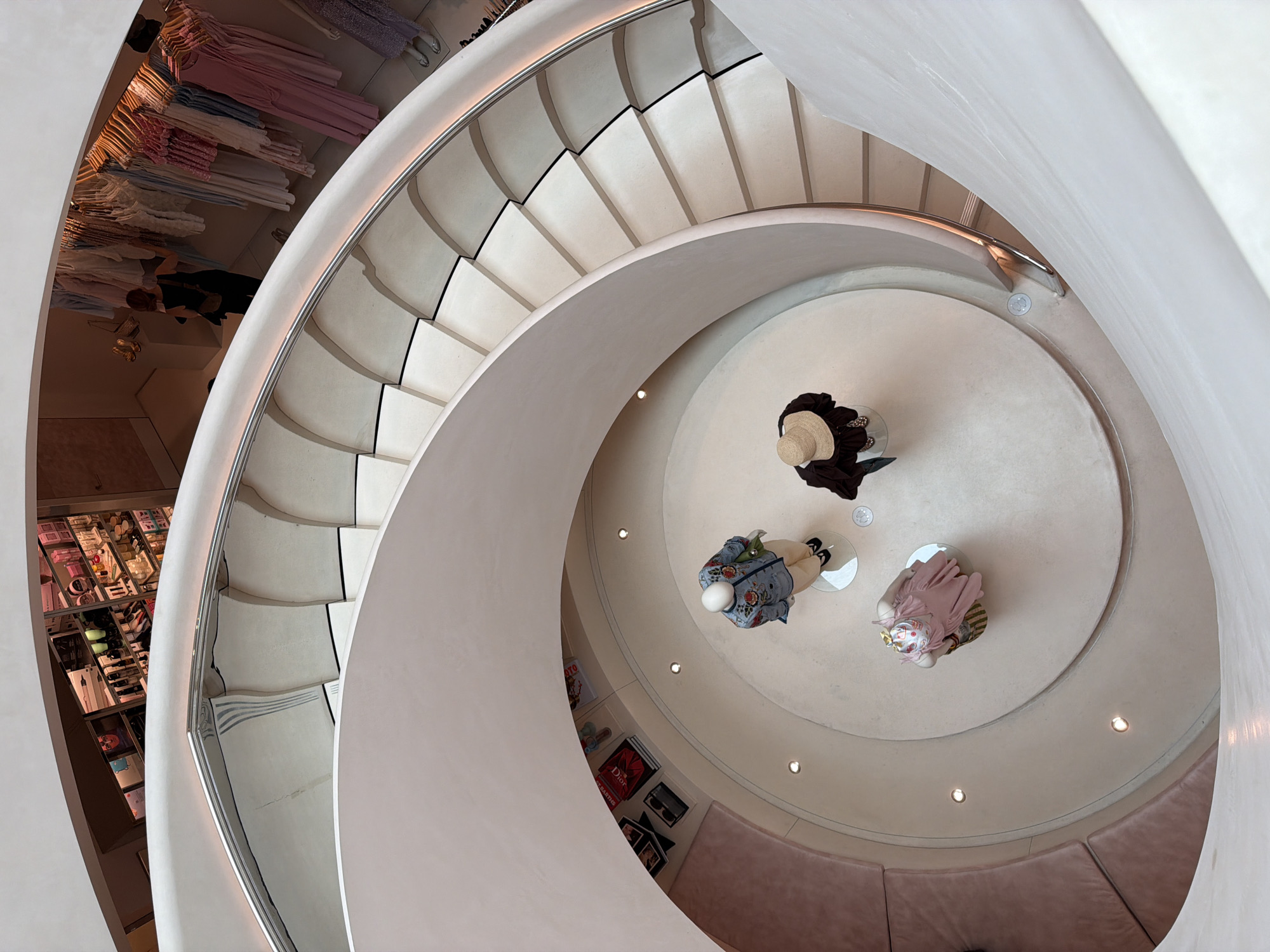 Viewed from directly overhead, a multi-level department store interior is dominated by a winding staircase. Mannequins stand at the center of the bottom floor. A seating area is curved and located in the interior of the bottom floor. A row of cosmetics lines a wall of the second floor. Clothing hangs on racks on the upper level. The photograph was taken inside the Reformation store in Melrose, Los Angeles, USA.