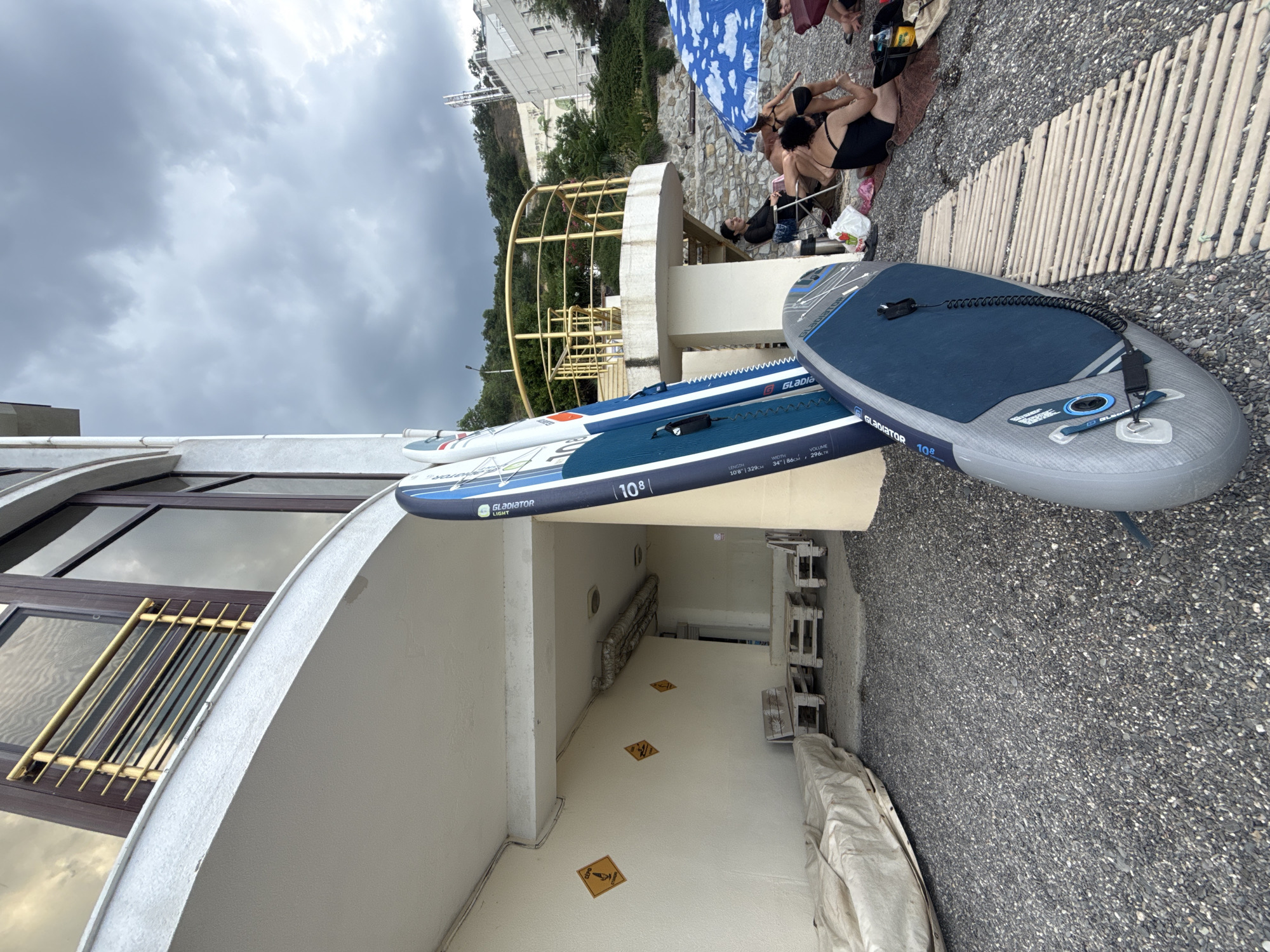 The image depicts an outdoor scene on a pebbly beach, likely in Alushta, Ukraine, with a building structure and several people in the background. In the foreground, three paddleboards are leaning against a cream-colored wall. The paddleboards are predominantly blue and white, with some branding visible. One paddleboard is lying flat on a wooden walkway made of thin slats.

To the left of the paddleboards, a large object covered in a light brown tarp is visible, positioned on the pebbly ground. The building behind the paddleboards has a white, curved overhang and large windows with dark frames. There are also yellow diamond-shaped warning signs attached to the wall. Further back, a yellow railing encloses a raised platform with stairs leading up.

In the midground and background, several people are seen sitting and interacting on the beach. One woman is seated in a folding chair under a blue and white patterned umbrella. Another person, partially obscured, is seated next to her. Some people appear to be wearing swimwear. Trees and a hillside covered in vegetation are visible beyond the building and railing, with a modern building structure on the far right. The sky is overcast with dark, cloudy conditions.