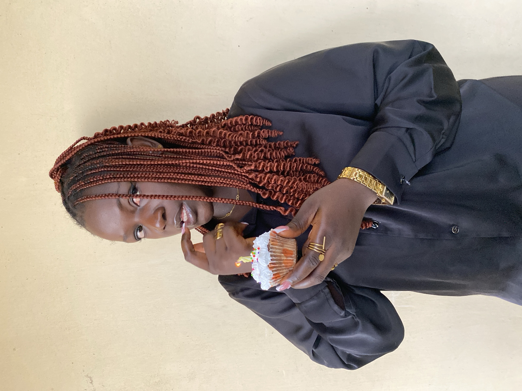A person with brown braided hair and dark skin is depicted indoors, holding a cupcake with white frosting and colorful sprinkles. The cupcake has a single lit candle. The person is wearing a black button-down shirt and gold jewelry, including a watch, rings, and a necklace. The background is a pale beige wall. The scene is located in Bukuru, Nigeria.
