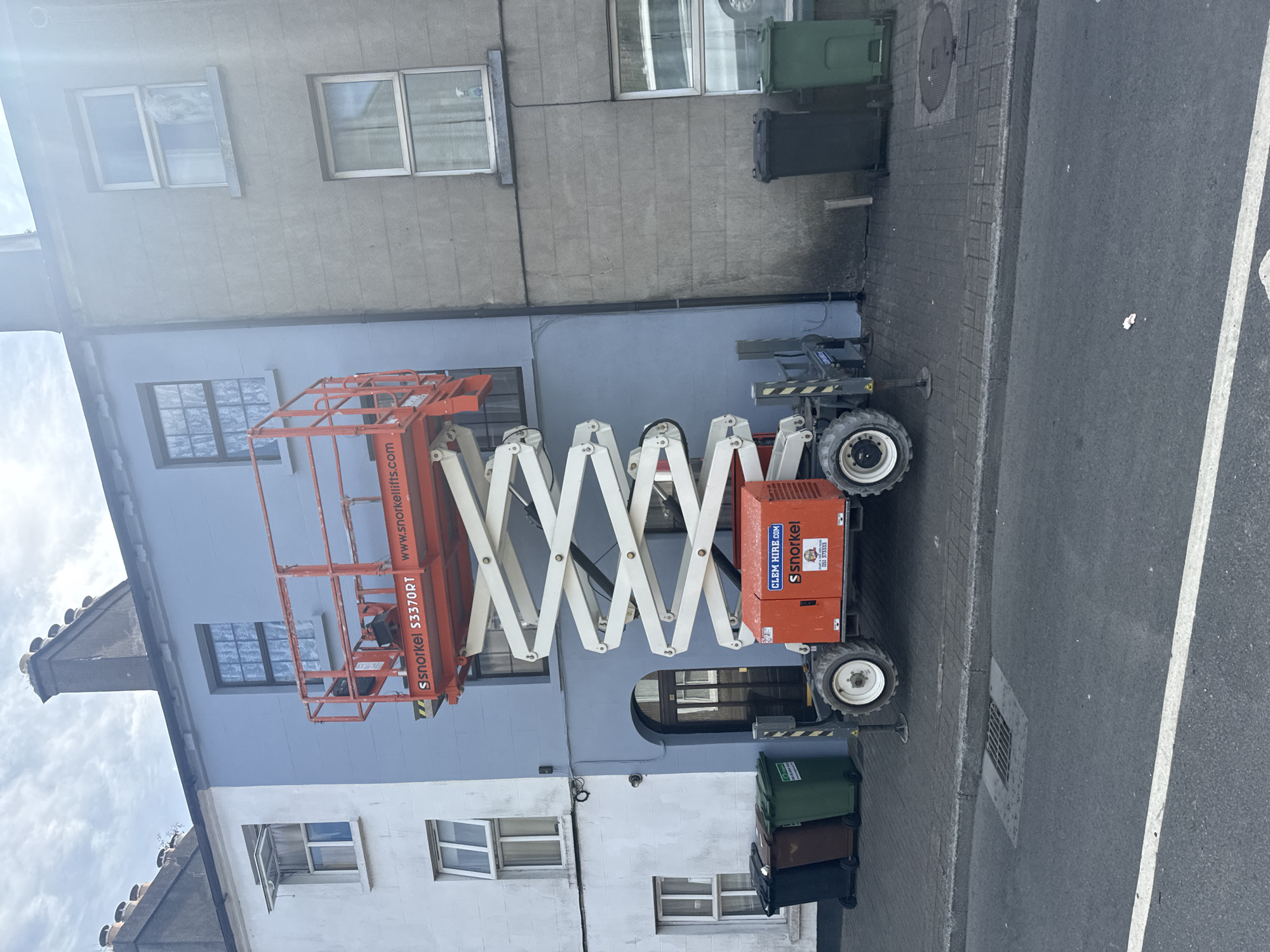 The image shows a Snorkel S3370RT scissor lift parked on a street in Waterford, Ireland, likely for building maintenance or construction. The scissor lift is positioned in front of a pale blue building, adjacent to a white building on the left and a grey building on the right. The platform of the scissor lift is extended upwards, with its orange railing visible against the blue wall. The base of the lift, which is orange, features branding that includes "SNORKEL" and "CLEM HIRE.com". Several residential buildings with windows are visible above the scissor lift. In the foreground, a street with a white line marking runs horizontally. Several trash bins, including black, brown, and green ones, are lined up on the sidewalk to the left of the scissor lift. The environment appears to be a typical urban streetscape.