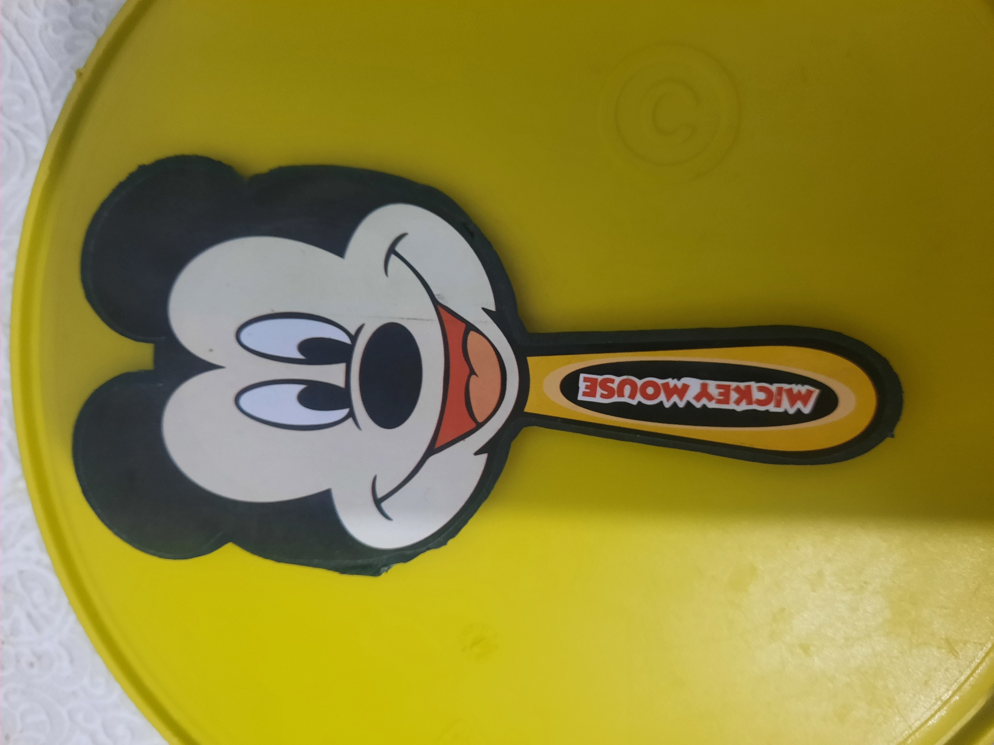 A Mickey Mouse-shaped hand mirror lies on a yellow surface. The mirror features Mickey Mouse's face with black ears, white eyes with black pupils, a black nose, a wide smiling mouth with red lips, and a small tongue. The handle of the mirror is black and yellow, with "MICKEY MOUSE" written vertically in white lettering on a red background. The yellow surface appears to be a plastic lid with a subtle circular embossed pattern on the right side. The background above the yellow surface is out of focus and shows a textured white surface. The image was taken in Lhokseumawe, Indonesia.