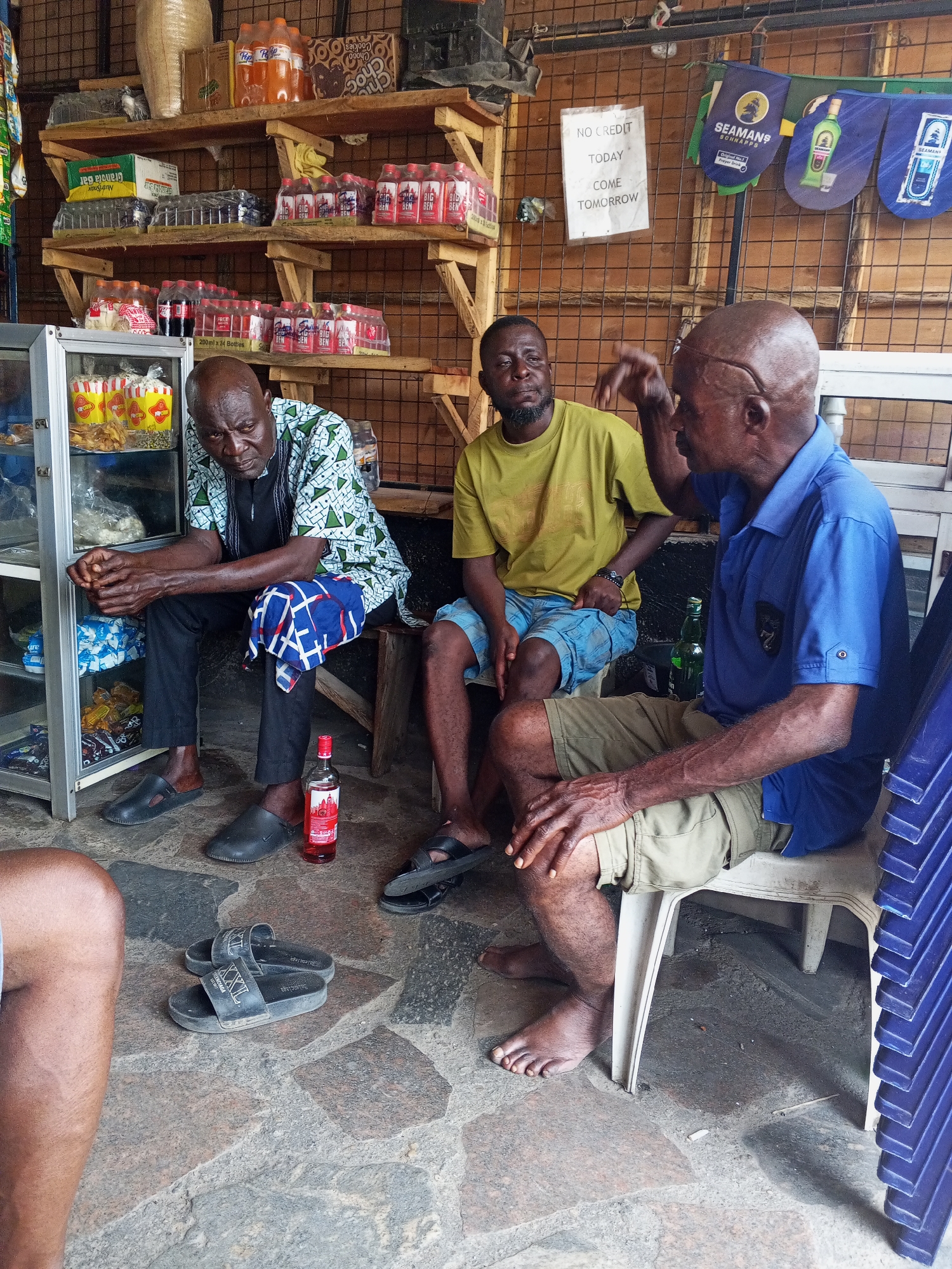 The image depicts three men sitting in a small shop or bar in Zango-ndaji, Nigeria. The man on the left wears a patterned shirt and black pants and sits next to a display case filled with snacks and drinks. A bottle of liquor sits on the floor next to him. The man in the middle wears a yellow t-shirt and blue shorts. The man on the right wears a blue polo shirt and khaki shorts and gestures with his right hand. A sign on the wall reads "NO CREDIT TODAY COME TOMORROW." Shelves behind the men are stocked with bottled drinks and snacks. Blue plastic chairs are stacked on the right side of the frame. The floor is stone.