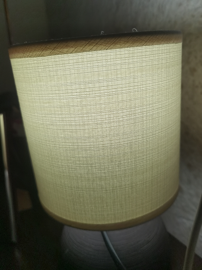 The scene, identified as being in Velichkovo, Bulgaria, depicts an indoor environment focused on a singular object. The central subject is an illuminated cylindrical lamp shade, fabricated from a light-toned, off-white or beige woven textile material exhibiting a consistent grid pattern. The upper and lower edges of the shade are bordered by a darker, possibly brown or golden-hued, trim. Below the shade, a segment of the lamp's dark, textured base is visible, from which a dark electrical cord descends, extending out of frame towards the lower left. The background is largely out of focus, comprising indistinct dark elements on the left, including a partially visible reflective vertical object, and a lighter, textured surface on the right. No human subjects or discernible activities are present.