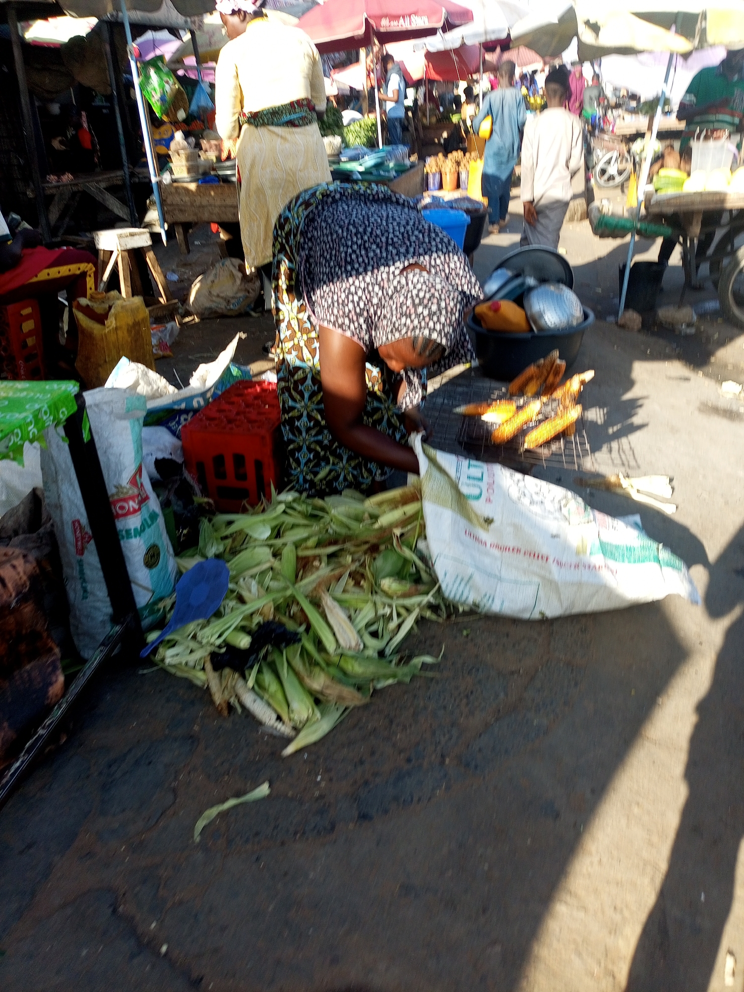This image depicts a busy street market scene in Tempure, Nigeria, with a focus on a food vendor grilling corn. The vendor, a woman wearing a patterned dress and a headscarf, is actively engaged in cooking the corn on a grill placed on the ground. Several ears of corn are visible on the grill, appearing to be roasted. In the foreground, there are large sacks and a pile of corn husks, suggesting fresh produce being prepared or sold. Scattered around the vendor are various items, including a blue scoop, a red crate, and cooking utensils in a black bucket. The background reveals more market stalls, umbrellas providing shade, and other people moving about, contributing to the bustling atmosphere of the marketplace. The ground appears to be a mixture of dirt and asphalt, typical of a street market environment. The sunlight casts strong shadows, indicating it is daytime.