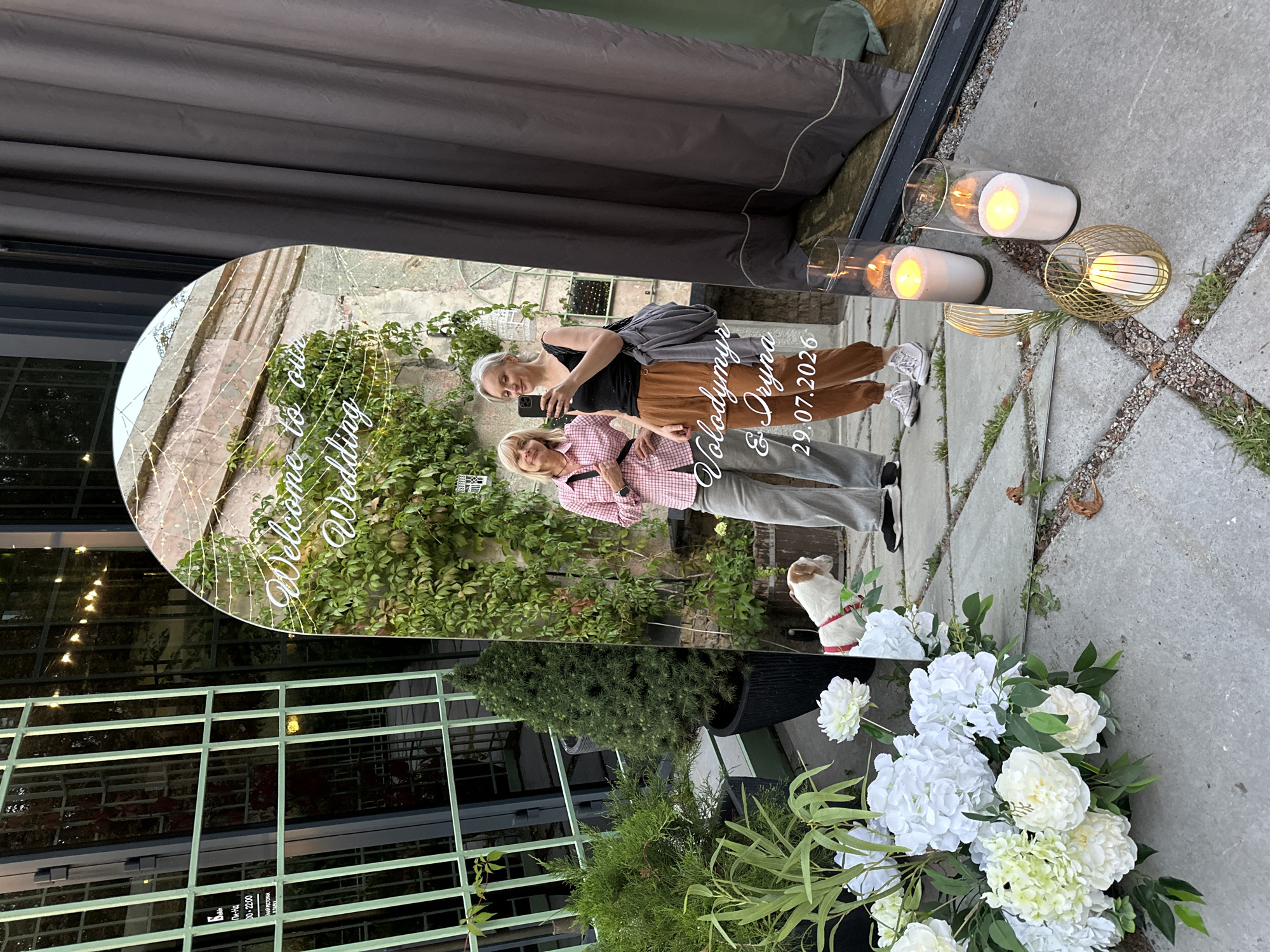This image depicts a wedding welcome mirror at an outdoor venue, possibly a cafe or restaurant in Kyiv, Ukraine. The mirror, which is arched at the top, has text etched onto its surface. The text reads "Welcome to our Wedding" at the top, followed by "Volodymyr & Iryna" and the date "29.07.2026" at the bottom.
Two women are reflected in the mirror. The woman on the left is older, wearing a pink and white checkered shirt and grey pants. The woman on the right is younger, dressed in a black tank top and brown pants, holding a smartphone and taking a selfie. A small white and brown dog, wearing a red leash, is standing on the pavement in front of the mirror.
To the left of the mirror, there are various potted plants, including a tall, conical evergreen tree and several large white flowers. Behind the plants is a building with a green metal grille facade. To the right of the mirror, a dark grey curtain hangs, and three glass vases with white candles and gold candle holders are placed on the stone-tiled ground. The background appears to be an old brick wall with ivy growing on it. The lighting suggests it is either late afternoon or early evening.