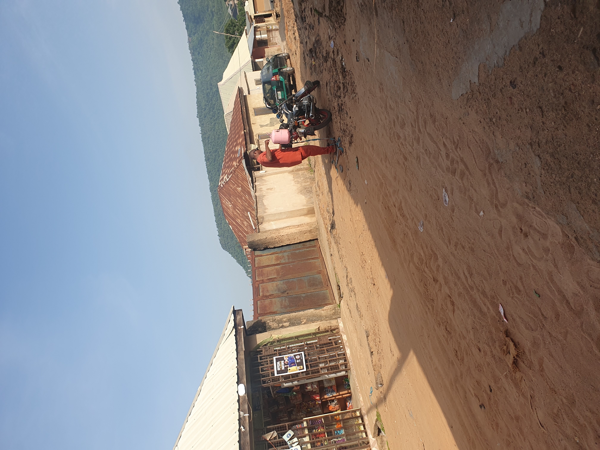 In the city of Lokoja, Nigeria, a man in an orange outfit is seen standing next to a motorcycle, holding a pink water container. The scene is a dusty, unpaved road with several buildings lining the sides. To the left, a small shop with shelves stocked with goods is visible, protected by a metal grate. Further down the road, a dark green SUV is parked. In the background, a lush, green hillside rises under a clear blue sky, suggesting a semi-urban or rural setting within Lokoja. The overall impression is one of a typical day in a Nigerian community, with elements of commerce and daily life.