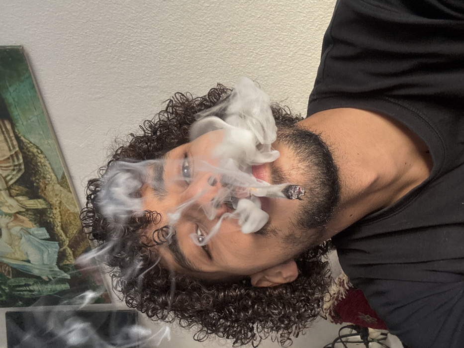 The scene, located in Tangier, Morocco, captures a close-up, eye-level view of a male individual indoors. The subject possesses dark, tightly curled hair and a short, dark beard. A silver ring is visible in a piercing on his right nostril. He is wearing a dark, crew-neck t-shirt. The individual is actively smoking a lit, hand-rolled cigarette, which is held between his lips. A substantial, dense cloud of white smoke billows from the cigarette and around his mouth and nose, partially obscuring the lower portion of his face. His eyes are open and directed forward, engaging the camera. In the background, a plain, light-colored wall is visible. Two pieces of art are affixed to the wall: an upper-right framed piece depicts classical figures in muted tones, and a partially visible, darker piece is situated further left. A section of patterned, woven fabric with red and cream colors is discernible in the lower-left corner of the frame.