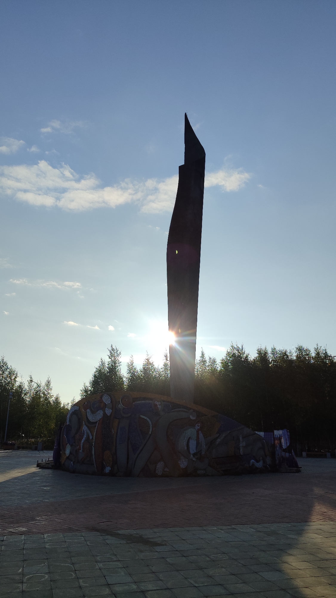 The image shows a monument in the city of Mirny, Russia. The monument is a tall, dark, abstract sculpture with a pointed top, silhouetted against a bright, sunlit sky. The sun is positioned behind the sculpture, creating a lens flare effect. The base of the monument is a large, curved structure adorned with colorful mosaic artwork depicting abstract figures and patterns. In the background, there is a line of trees, and further back, a clear blue sky with scattered white clouds. The foreground consists of a paved plaza with square tiles. The overall scene suggests an outdoor public space.