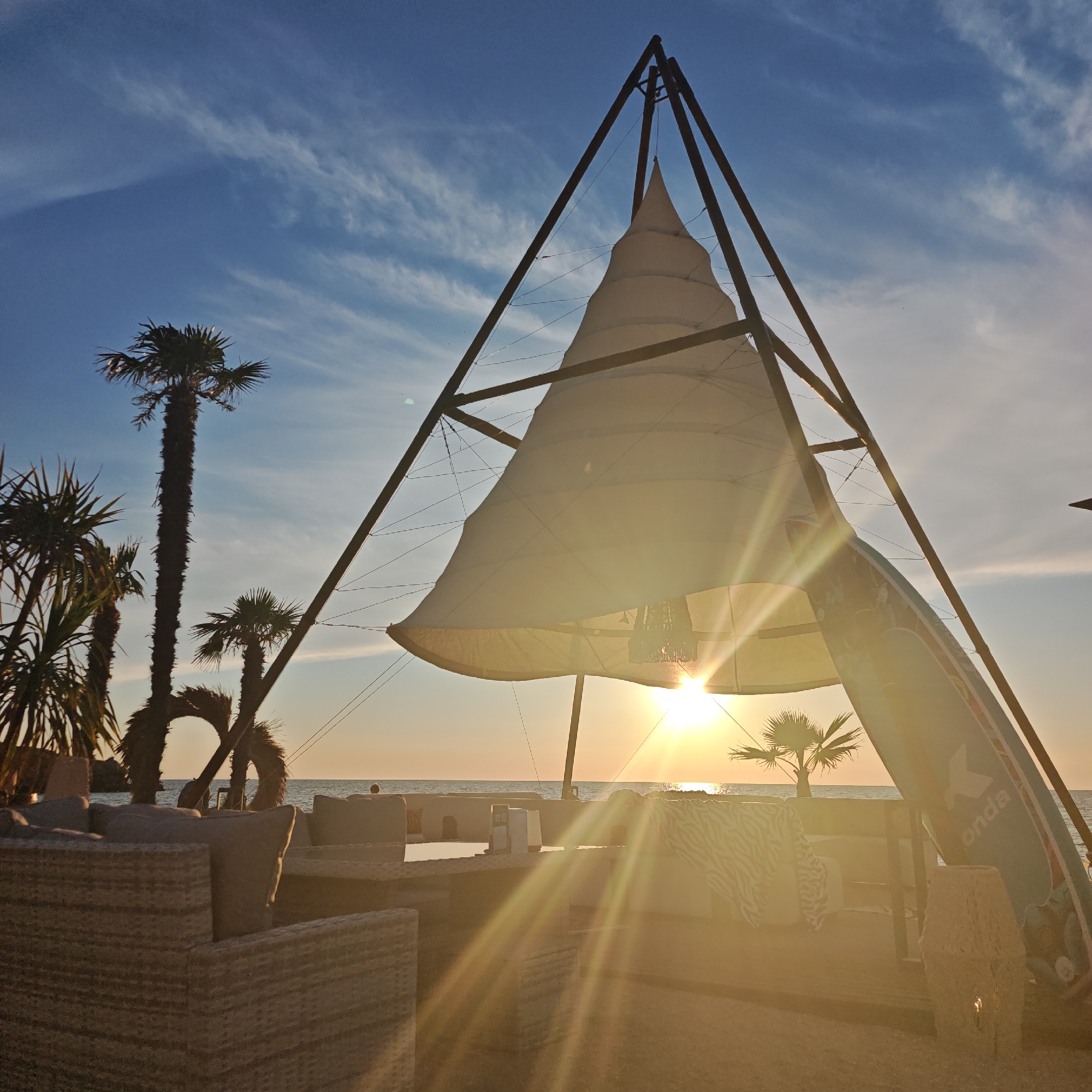 The image depicts a sunset scene at a beachside cafe or resort in Мысовское сельское поселение, Ukraine. The foreground features outdoor seating made of wicker furniture with plush cushions. Behind the seating, a large, conical canvas structure supported by a metal frame is visible, serving as a shade or canopy. The sun is setting, casting a warm, golden light with prominent lens flares across the scene, illuminating the canvas and the ocean in the background. Several palm trees are present, adding to the tropical ambiance. A surfboard is leaning against the structure on the right side of the image, further suggesting a beach or water-related setting. The sky is a clear blue with wispy white clouds, transitioning to a warmer hue near the horizon. There are no people visible in the image.