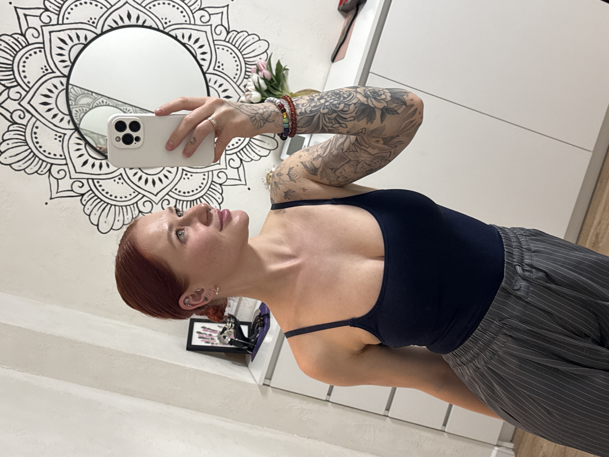A fair-skinned woman with auburn hair takes a mirror selfie. She wears a dark blue tank top and gray pinstriped pants. Her left arm is raised, holding a white smartphone, revealing tattoos on her arm. She has multiple ear piercings. A round mirror with a mandala design surrounds it is mounted on the wall behind her. To the left, a black framed picture sits on a white shelf. A bouquet of tulips is also present in the background. The setting appears to be indoors, possibly in a bedroom or dressing area with white walls and cabinets. There is no evidence of a location in Rostov-on-Don, Russia.