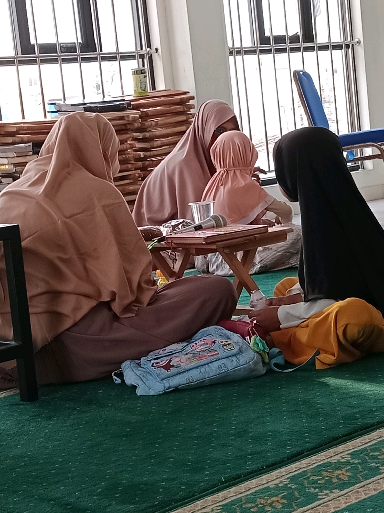 The image depicts a group of individuals, likely children and adults, engaged in an activity indoors, possibly a religious or educational setting. They are seated on a green carpeted floor in front of a large window with metal bars. To the left, a stack of wooden planks is visible. In the center, a small wooden table holds a book, a microphone, and a plastic cup. A blue backpack with cartoon characters is placed on the floor in the foreground. The people are wearing headscarves, and their attire suggests a cultural context where such clothing is common. The environment appears to be a room with plain walls and a simple interior. The overall scene suggests a moment of learning or communal gathering.