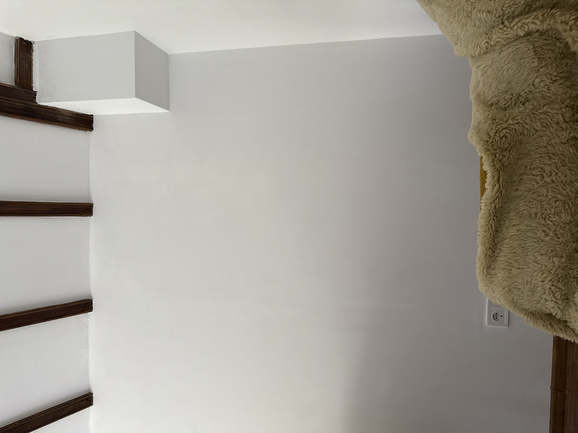 The interior of a room in Razgrad, Bulgaria features white walls, with dark wooden beams creating a decorative element on the left side. A white box-like architectural detail is positioned at the top of the wall on the left. On the right side, a large, shaggy, beige fabric partially obscures the wall. An electrical outlet is visible on the lower right side of the wall, partially covered by the fabric. The lighting is even and diffuse.