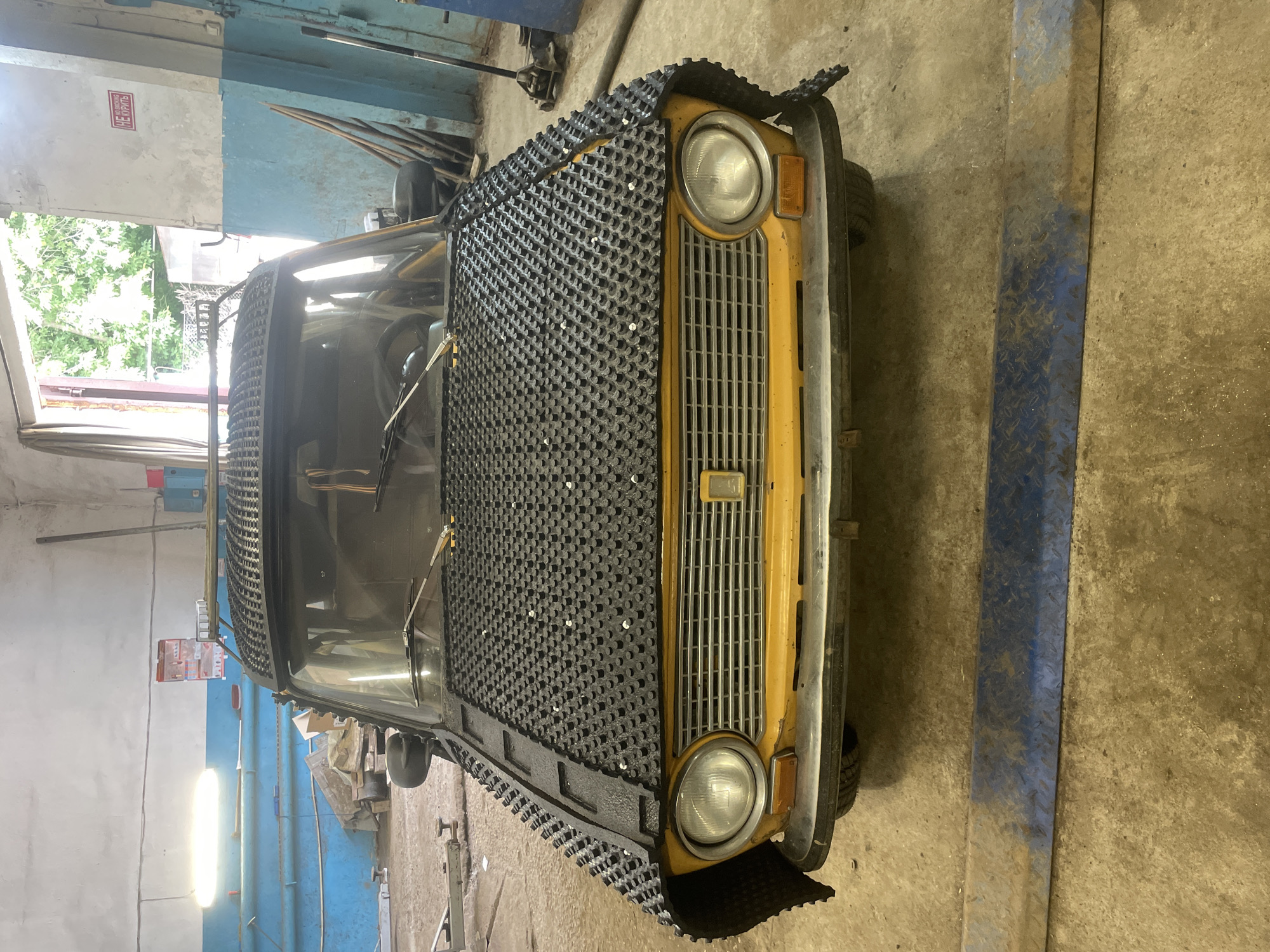 The image shows a yellow car with a unique black grille cover. The car is parked in what appears to be a garage or workshop. The grille cover is made of a textured material with a grid pattern and appears to be custom-fitted to the front of the vehicle, covering the headlights and grille.
The setting is a dusty concrete floor, with blue and light-colored walls in the background. There are some tools and equipment visible, including a car jack and what looks like metal shelving. Natural light seems to be coming from an open doorway or window on the left, revealing some greenery outside.
A red and white sign with Cyrillic text is mounted on the wall to the left of the car. The text on the sign reads "НЕ БОИРЬ", which translates to "Don't be afraid".
The image is a static shot, with no activity clearly depicted, other than the car being present in the workshop. The lighting suggests it could be daytime. The overall impression is one of a utilitarian space where a vehicle might be undergoing maintenance or modification.