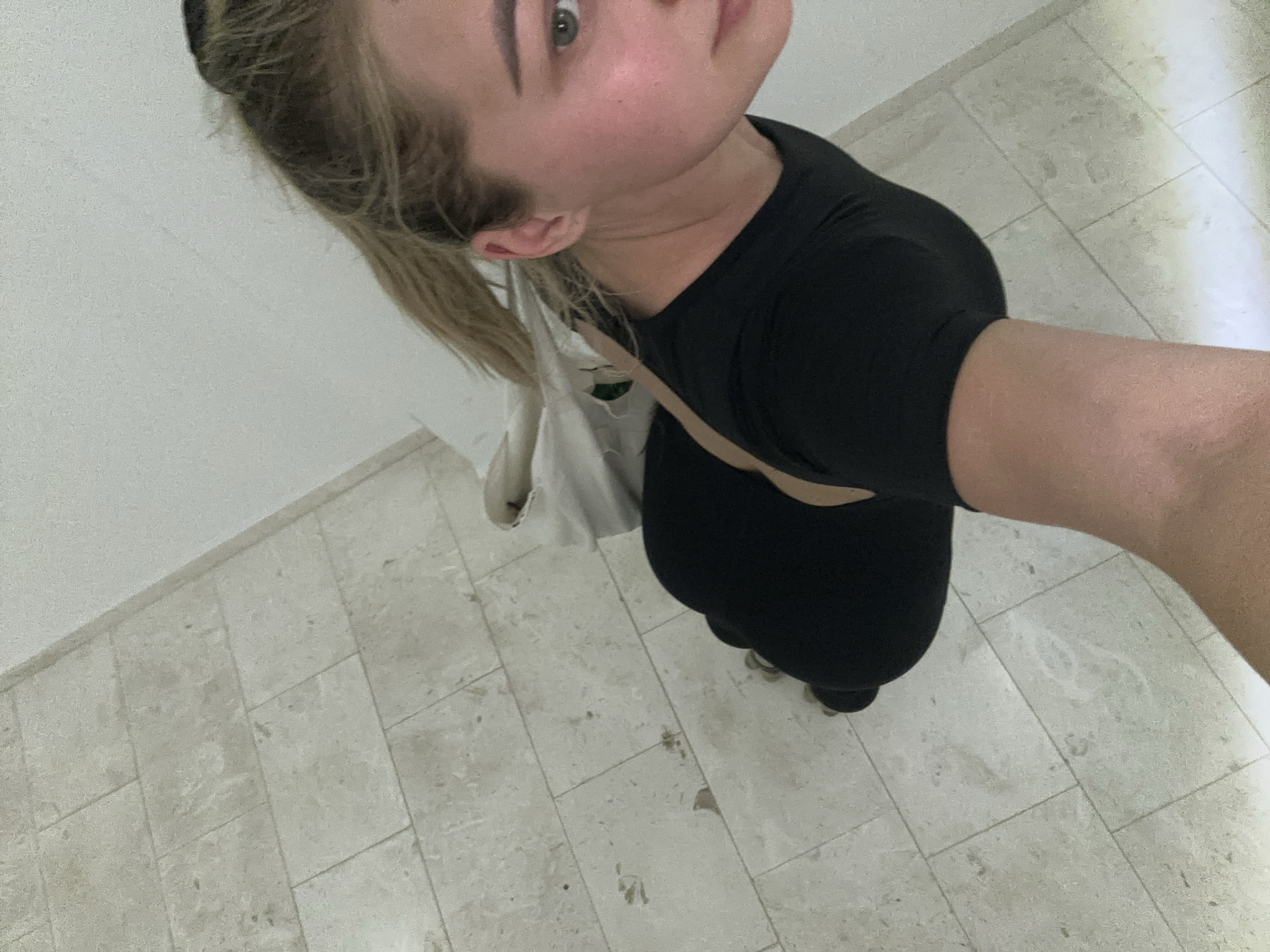The image is a high-angle, downward-looking selfie taken indoors by a person, likely female, in Budva, Montenegro. The subject has light-colored hair, pulled back into a ponytail, and light eyes, looking up towards the camera with a neutral expression. She is wearing a black, form-fitting, short-sleeved top or dress. A light-colored (possibly white or off-white) tote bag is slung over her left shoulder or arm.
The setting consists of plain, light-colored walls and a light-toned tiled floor. The rectangular floor tiles are arranged in a grid pattern and show some subtle variations in color and texture, with a few small dark marks. The person's body is positioned diagonally across the frame, with her head and upper torso in the upper left, and her body extending towards the lower right, with her right arm (presumably holding the camera) visible on the far right. The lighting is bright and even, suggesting it is daytime. No discernible text is visible on the subject's clothing, bag, or the surrounding environment. The scene depicts a casual, everyday moment.