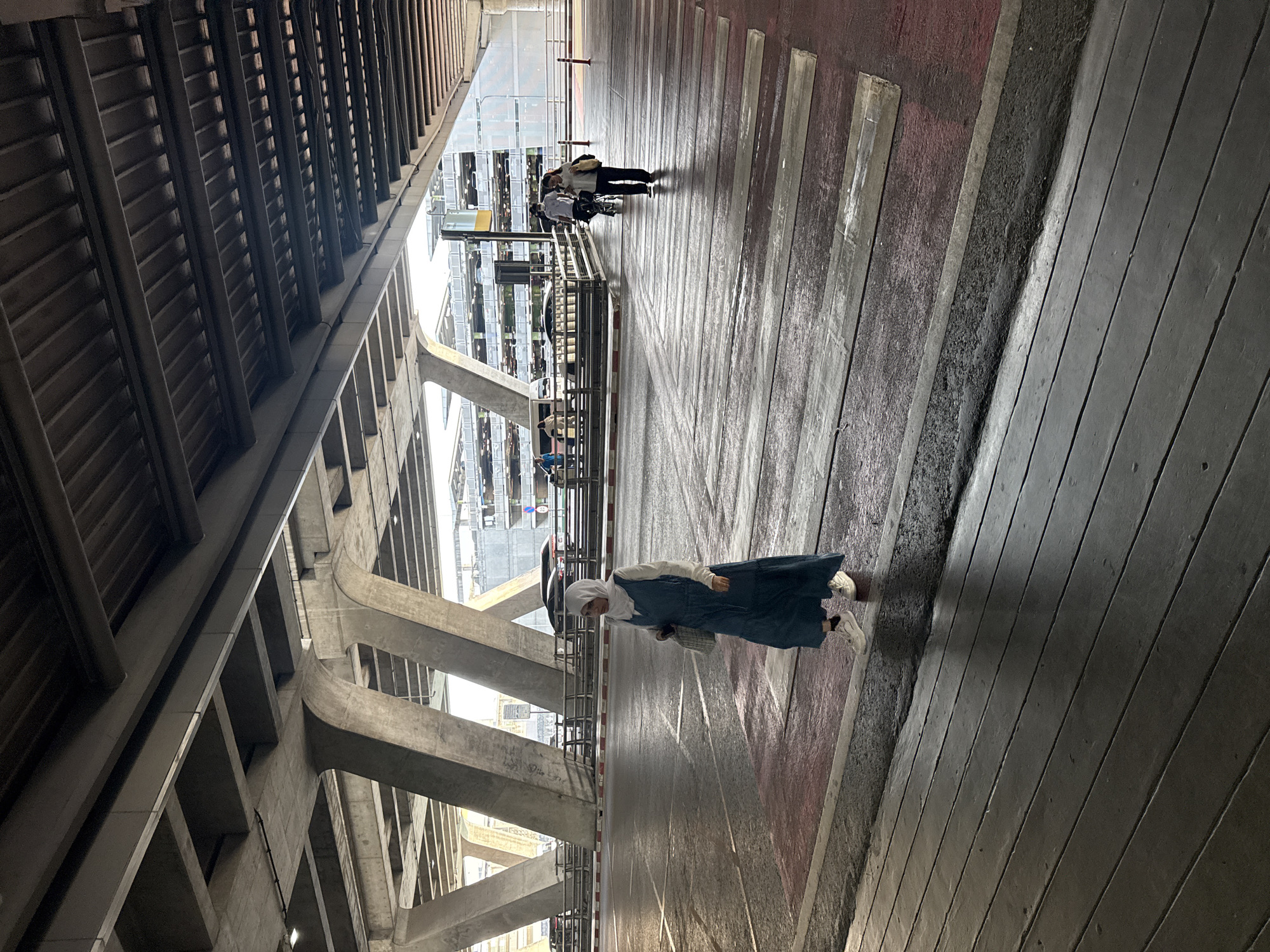 A full-shot image shows a person walking across a pedestrian crossing on a wet asphalt road. The person, wearing a blue dress and a white hijab, walks from left to right towards the center of the frame. They are carrying a bag. Behind them, two more individuals are walking on the crossing from right to left. The foreground shows a wooden walkway. The background is a multi-level parking structure. Concrete pillars and a ceiling structure are visible above, casting shadows on the scene. The image appears to be taken in an urban environment, likely in Nong Prue Subdistrict, Thailand, given the context.