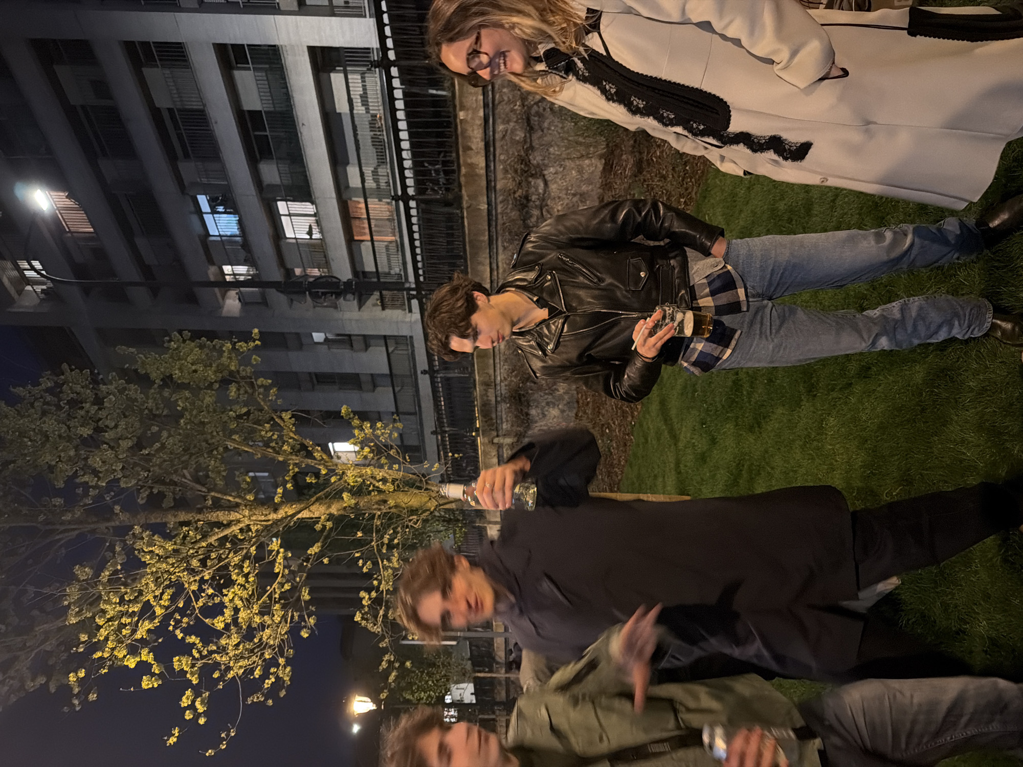 The scene is an outdoor gathering at night. A woman in a white coat with black trim is standing on grass, alongside a man in a black leather jacket, who is holding a glass. A third man in a black coat is pouring from a bottle into another glass held by a fourth man in a green jacket. A tree with blooming flowers is in the background and a tall building is in the distance. The setting is likely in Paris, France, based on the architecture and street lighting.