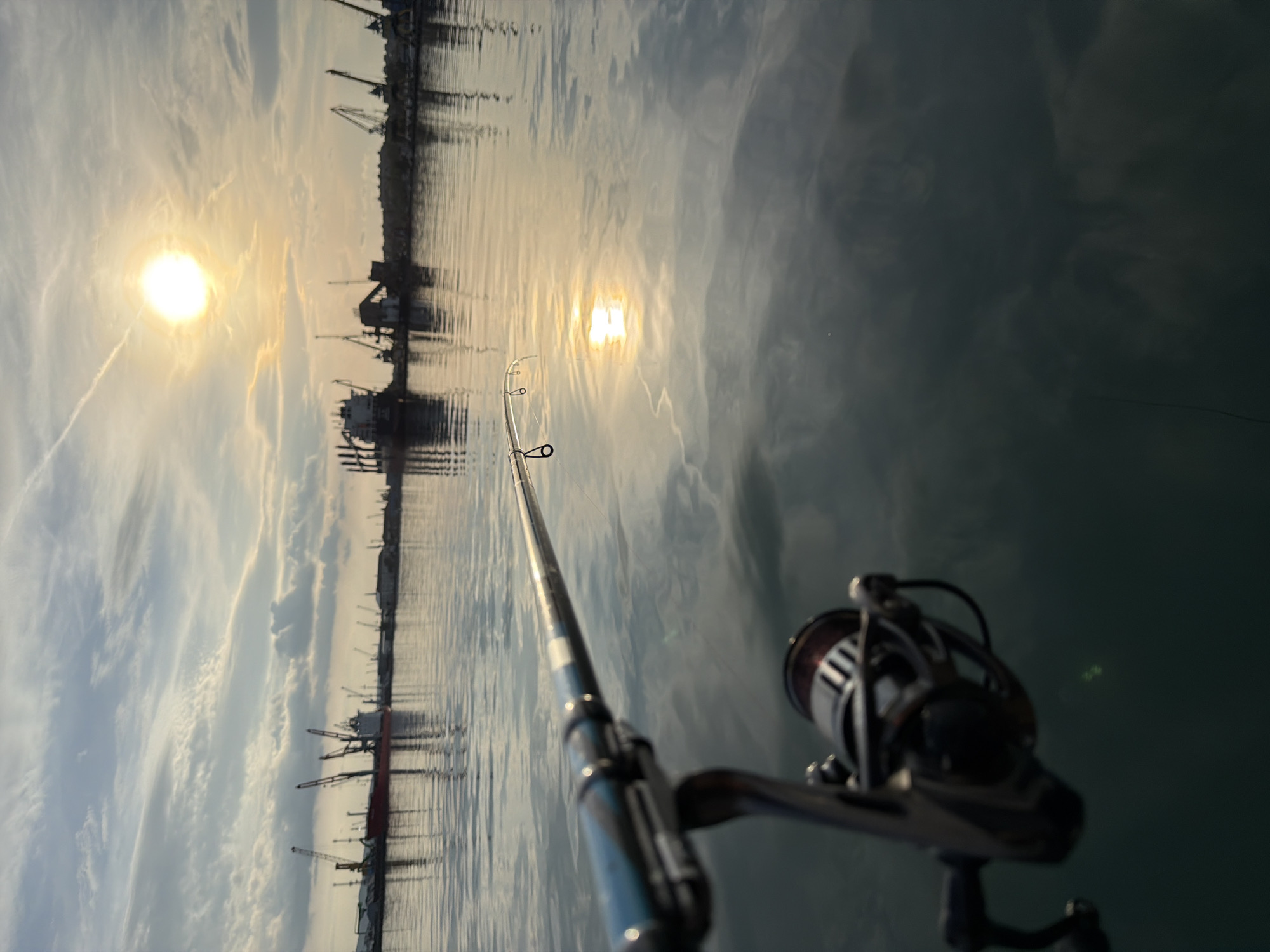 The image shows a fishing rod pointing towards the water, with the sun and reflections visible on the surface. In the background, there are port structures, including cranes and buildings, likely located in the city of Burgas, Bulgaria, as the city is known for its port. The fishing rod and reel are in the foreground, suggesting the perspective of someone fishing. The time of day appears to be either sunrise or sunset due to the position of the sun. The water is calm.