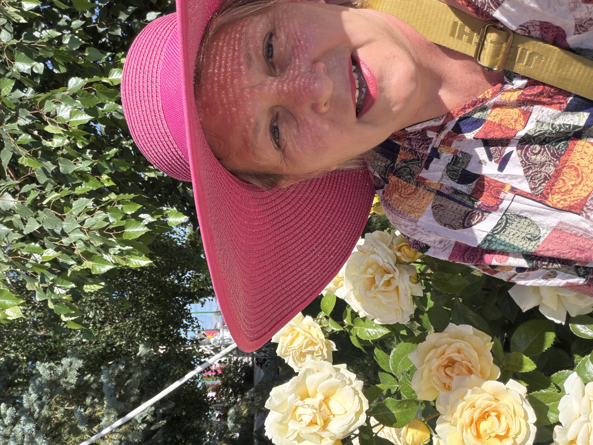 This is a portrait-style, close-up, over-the-shoulder shot of a woman smiling and looking towards the camera. She is wearing a wide-brimmed, bright pink straw hat that casts a patterned shadow across her face. She has blonde hair and is wearing bright pink lipstick. Her clothing is a colorful, patterned blouse with a yellow strap across her chest. Behind her, a bush with light yellow roses is visible, as well as green foliage from trees and a hint of blue sky. The lighting suggests it is a sunny day, possibly mid-day due to the strong shadows and bright illumination. The scene appears to be outdoors, perhaps in a garden or park, given the presence of flowers and trees. There is no visible text in the image. The woman's expression and the bright colors suggest a cheerful and pleasant mood.