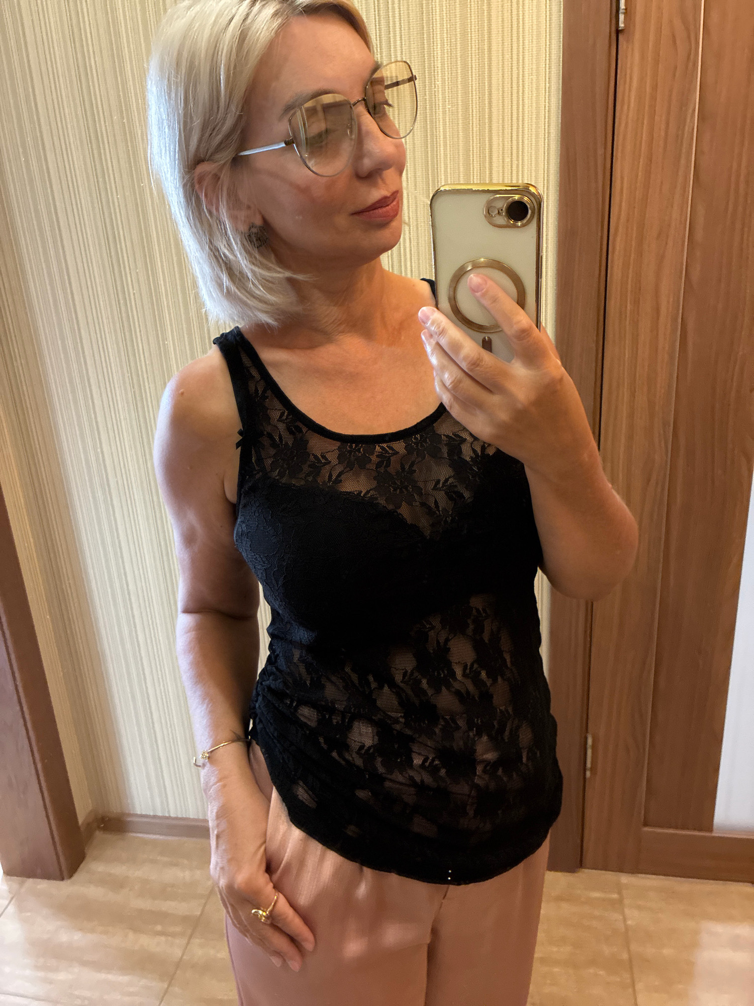 The image shows a woman with blonde hair and glasses taking a selfie in a room. She is wearing a black lace top and pink pants. The background consists of a wall with vertical stripes and a wooden door. The lighting suggests an indoor environment. There are no specific details in the image that would indicate the location as Sochi, Russia.