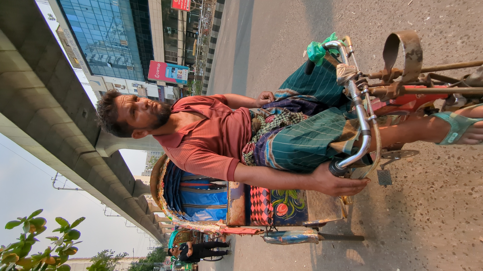 A rickshaw puller sits idle on his vehicle along a city street in Dhaka, Bangladesh. The man has a beard and dark hair and is wearing a rust-colored, short-sleeved shirt and a green and blue checkered lungi. He is holding onto the rickshaw's handlebars and has his feet on the footrest. The rickshaw's carriage is blue with patterned fabric and painted decorations. The street is asphalt, and in the background, a modern glass building is visible with a bridge or elevated structure extending over the road. A man in black stands near the rickshaw. A sign with an advertisement is also visible in the background.
