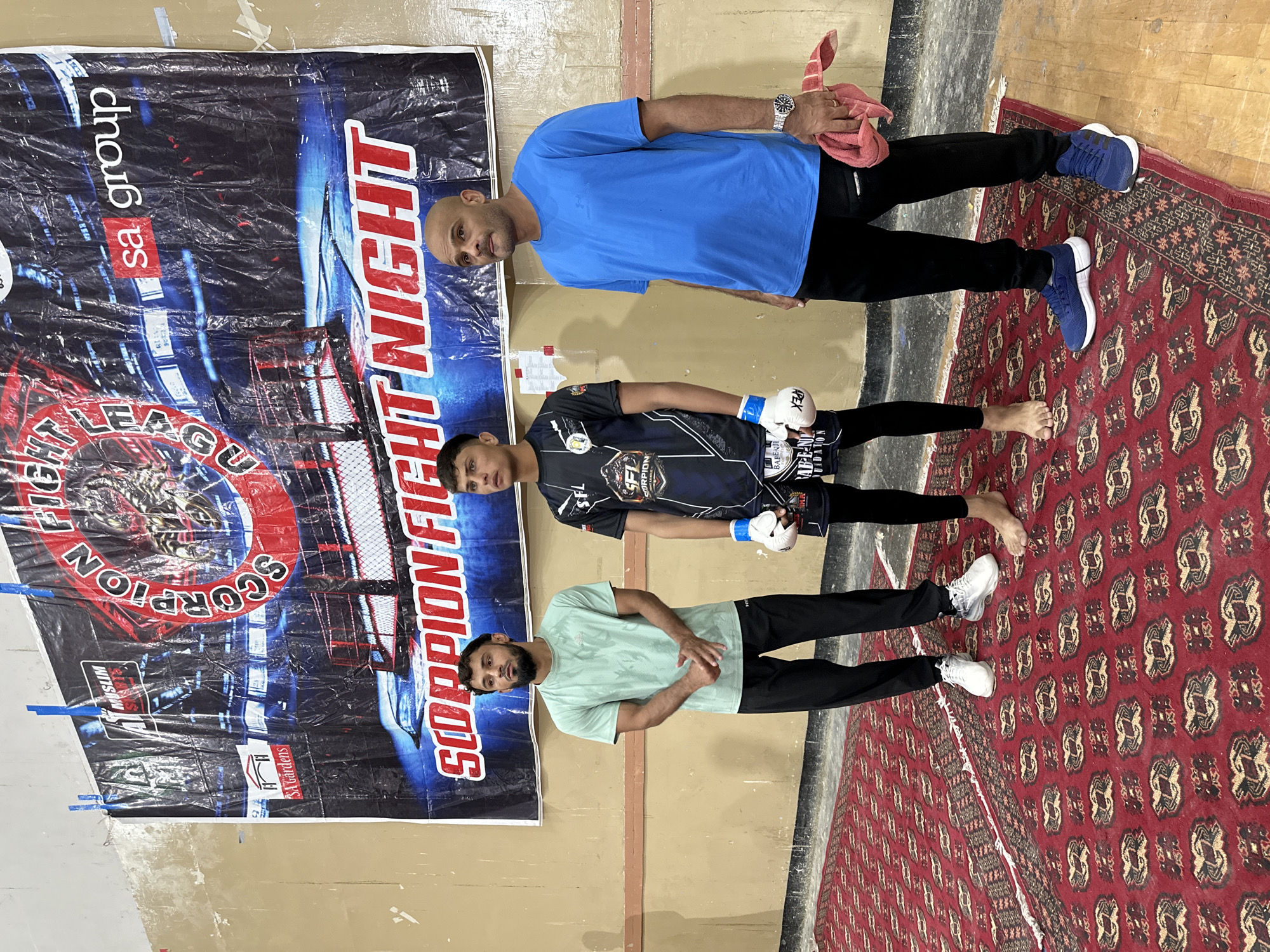Three adult males are standing in a room. The man in the center is wearing boxing gloves. Behind them is a banner that reads "SCORPION FIGHT NIGHT" and "sa group". The floor is covered with a red patterned rug. The location is Quetta City Tehsil, Pakistan.