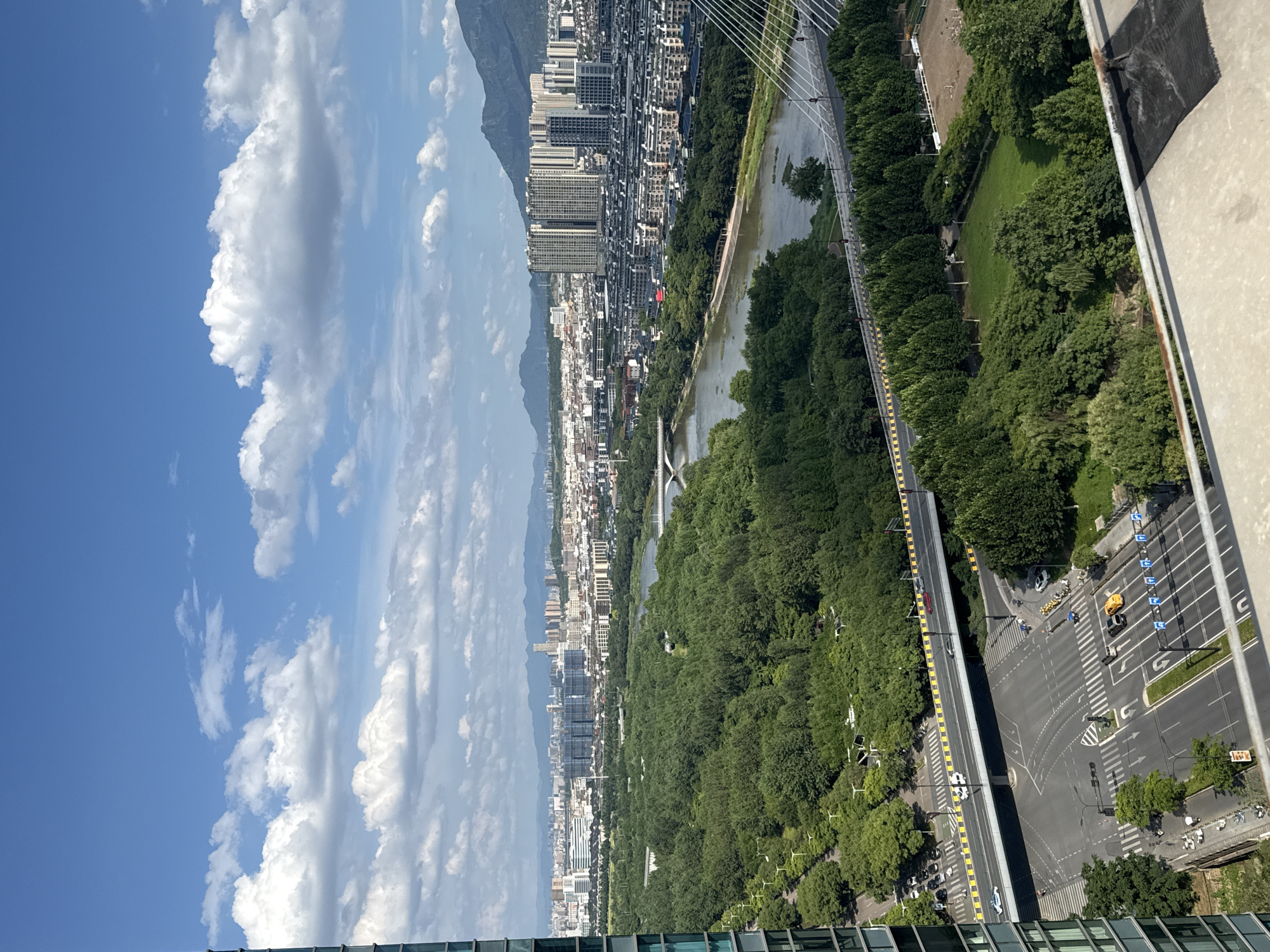 This is an aerial view of Yiwu, China, showing a cityscape dominated by buildings, a river, a bridge, and a large park or forested area. The sky is blue with scattered white clouds. In the foreground, a tall glass building is partially visible on the left. Below, a multi-lane road intersects with another road, with several cars, motorcycles, and a yellow taxi visible. A pedestrian bridge crosses over the intersection. Beyond the intersection, a wide, tree-lined avenue leads towards a large green space. A river flows through the middle ground, spanned by a modern suspension bridge on the right and a smaller bridge further left. The riverbanks are lined with trees. In the background, a dense urban area with numerous apartment buildings and taller structures stretches towards a range of mountains under the hazy sky. The overall scene depicts a blend of urban development and natural green spaces within the city.