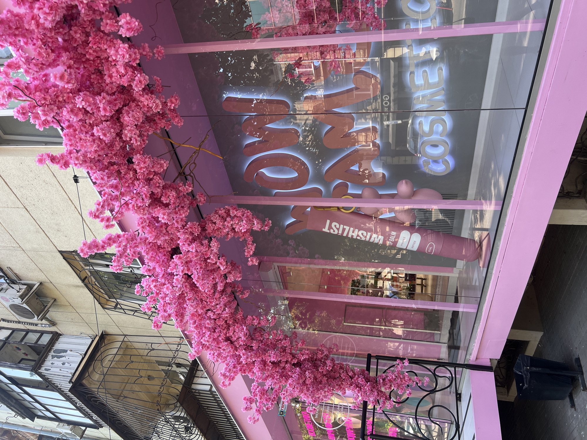 The image shows the exterior of a pink cosmetics store in Odesa, Ukraine, decorated with a large artificial cherry blossom arch. The store has large glass windows, one of which displays a neon sign that reads "WOW PAM COSMETIC" in large, stylized letters. A large pink tube, possibly a product display, stands near the window with the text "WISHLIST LIP BALM" visible. Pink balloons are also visible inside the store. Outside the store, a black trash bin stands on the pavement. In the background, a building with balconies and air conditioning units is visible. The scene appears to be a street-level view of a commercial establishment in an urban environment.