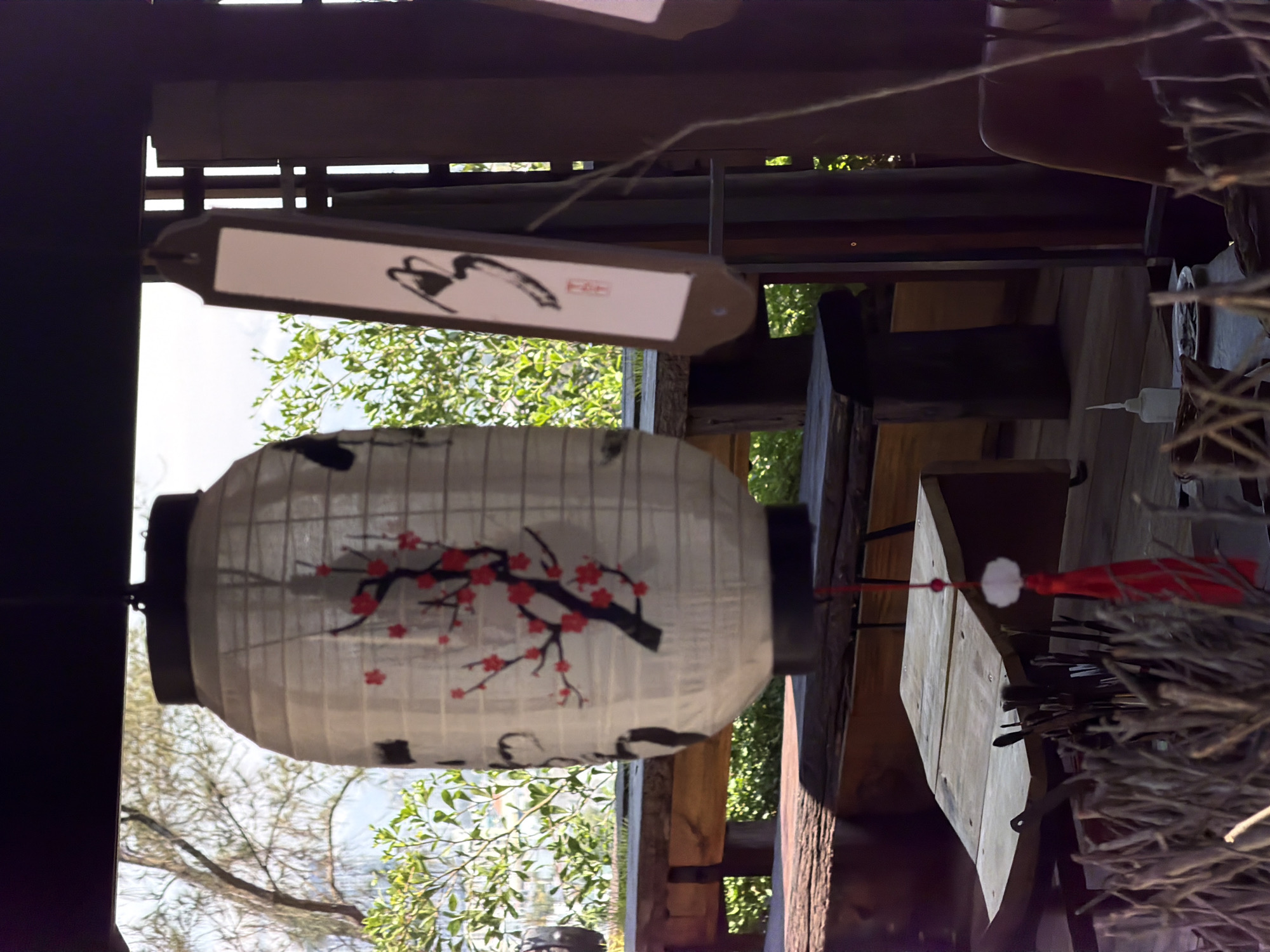 A daytime shot from Đà Nẵng, Vietnam, shows a white, spherical paper lantern with black trim and a painted design of a black branch bearing red flowers. The lantern hangs from a wooden structure. Above the lantern, there is a rectangular wooden sign with painted East Asian characters and a red stamp. The surrounding environment includes green foliage and a glimpse of the sky. A wooden table and other wooden structures are partially visible to the right of the lantern. Red tassels hang from a wooden surface to the right. Dried branches are in the foreground of the shot to the right.