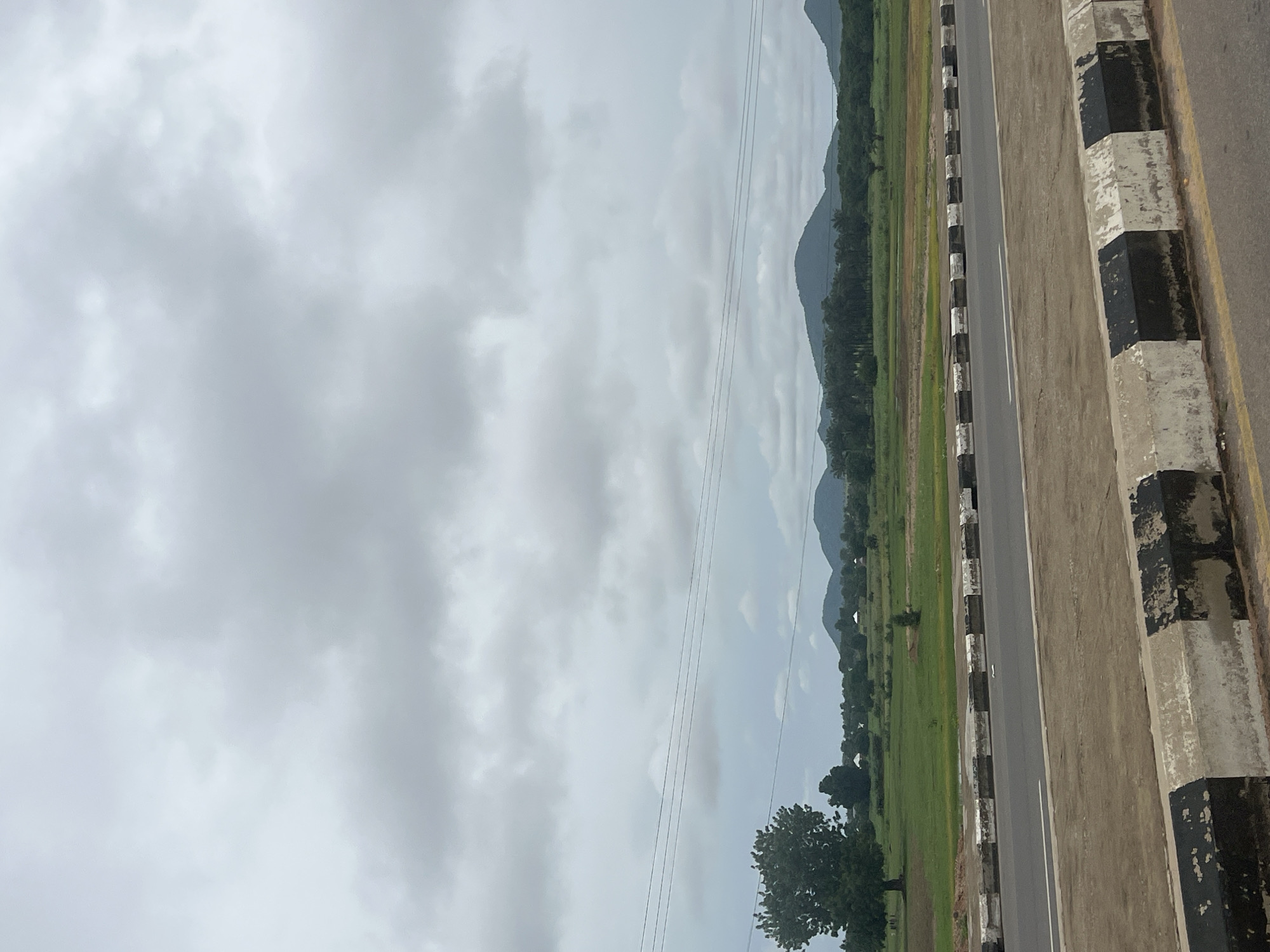 The image depicts a wide, paved road with a distinctive black and white striped barrier along its edge. Beyond the road, a rural landscape unfolds under a cloudy sky. A large, solitary tree stands prominently on the left, with green fields stretching out behind it. In the distance, rolling hills covered in vegetation form a backdrop, with several rounded mountain peaks visible. The area appears to be semi-arid, with patches of dry grass interspersed with greener areas. Several small, white-roofed structures, possibly homes or small buildings, are scattered across the landscape. The overall impression is one of a tranquil, undeveloped area on the outskirts of a city.