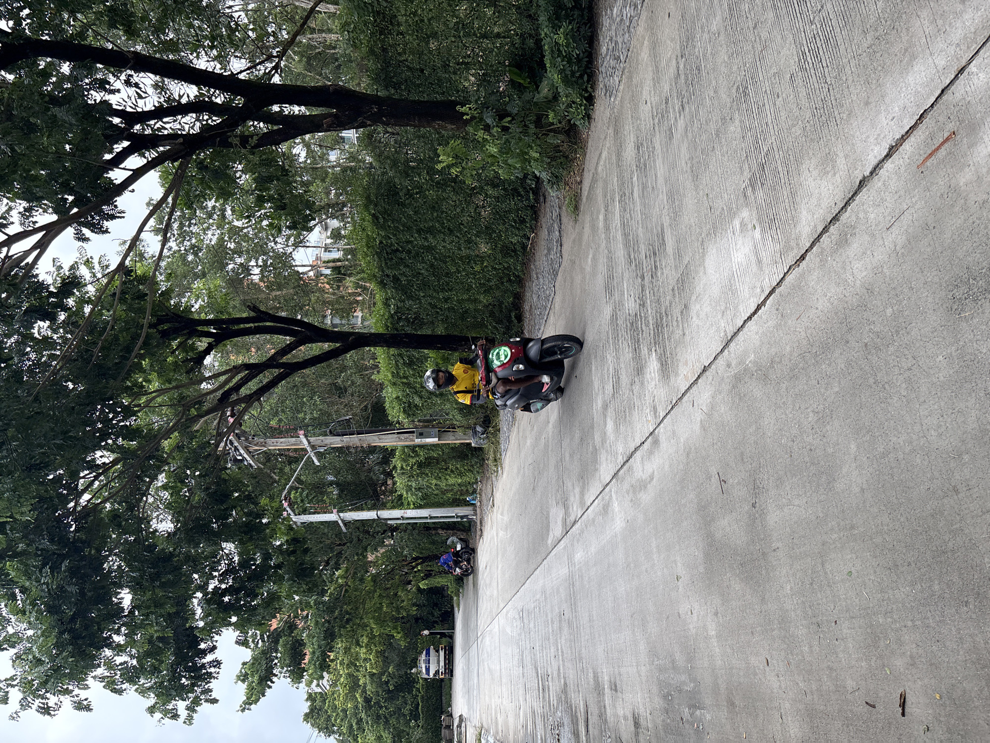 In Sakhu, Thailand, a person wearing a yellow shirt and a helmet rides a dark-colored scooter with red accents on a wet concrete road. The scooter's headlight is illuminated. The road is flanked by lush green trees and hedges, with a clear sky visible through the branches. In the distance, a white truck with a blue tank and another person on a motorcycle can be seen. The scene appears to be in a residential or semi-rural area.