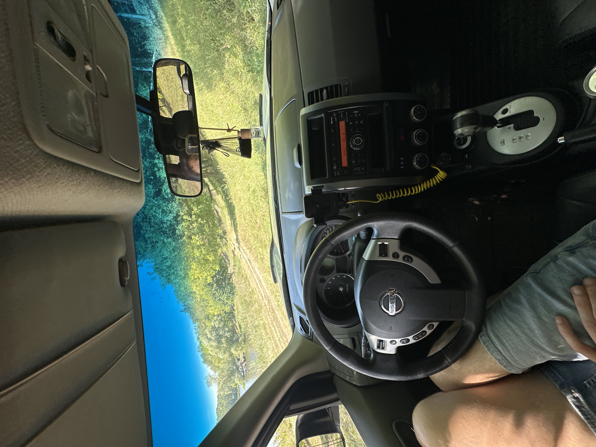 An interior shot of a Nissan vehicle shows a dashboard, steering wheel, and center console. Outside the windshield, a rural landscape with a road, trees, and a body of water is visible under a bright blue sky. A person's legs, wearing denim shorts and a t-shirt, are in the lower left corner of the frame. A car air freshener hangs from the rearview mirror. The image appears to have been taken on a sunny day. The location is Makiv, Ukraine.