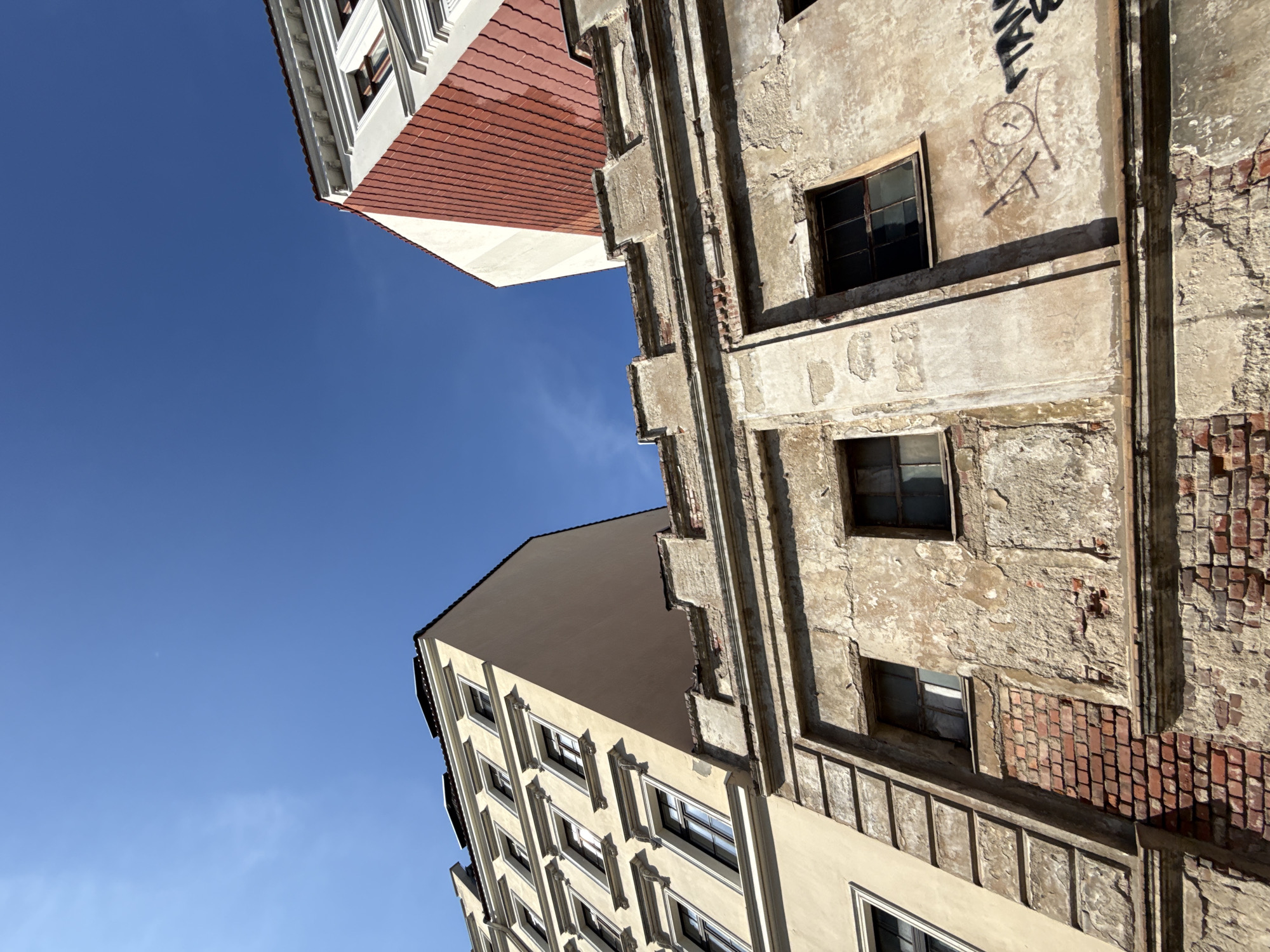 The image shows a view looking up at the facades of old buildings against a clear blue sky in Görlitz, Germany. In the foreground, a weathered, two-story building with exposed brickwork and peeling plaster is visible. It features several small, dark-paned windows. Above this, a taller, pale yellow building with multiple windows, each framed by decorative molding, rises. To the right, a portion of another building with a red-tiled roof and white and red painted walls is visible. Graffiti is present on the wall of the foreground building. The perspective is from a low angle, emphasizing the height and architecture of the buildings.