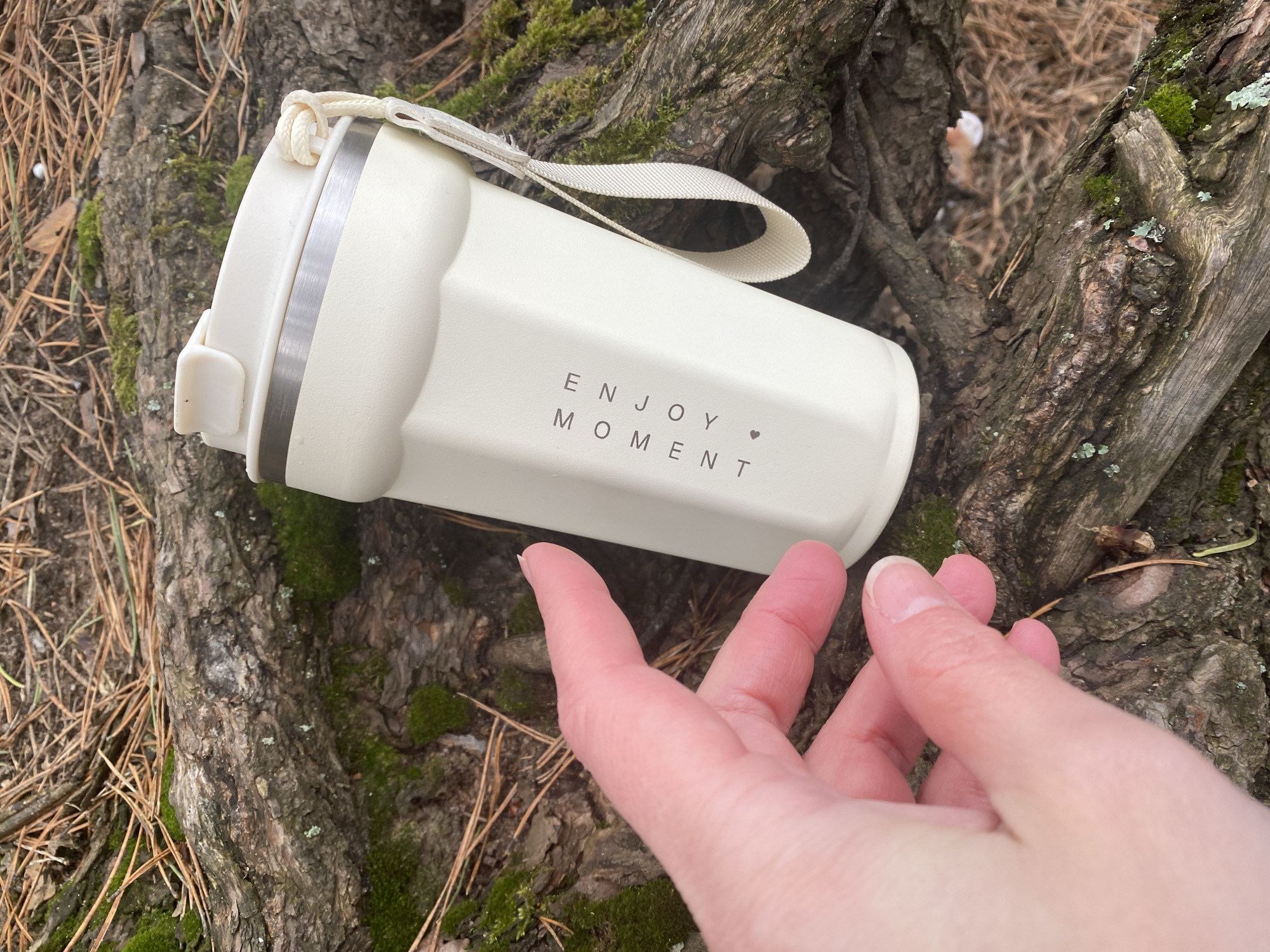 The image depicts an outdoor scene in Chaikovsky, Russia, focusing on a cream-colored tumbler resting on the prominent roots of a large tree. A light-skinned human hand, with fingers gently splayed, is positioned near the tumbler, suggesting interaction or an impending grasp.
The setting is a natural, wooded environment, characterized by the textured, dark brown tree roots and bark, which are partially covered with patches of green moss and some pale lichen. The ground surrounding the roots is strewn with dry, brown pine needles, indicative of a coniferous forest.
The tumbler, the central object, is off-white or cream, featuring a subtle, possibly ribbed texture on its body and a sleek metallic band near the top. A white fabric strap or handle is attached to the lid, which also has a small, light-colored plastic component. Clearly visible on the tumbler's side is the phrase "ENJOY MOMENT" printed in capitalized, spaced-out letters, followed by a small heart icon.
The scene conveys a sense of tranquility and a connection with nature. The soft, natural lighting suggests it is daytime, possibly overcast or within the shade of trees. The overall impression is one of a peaceful pause or an appreciation for the natural surroundings, emphasized by the message on the tumbler.