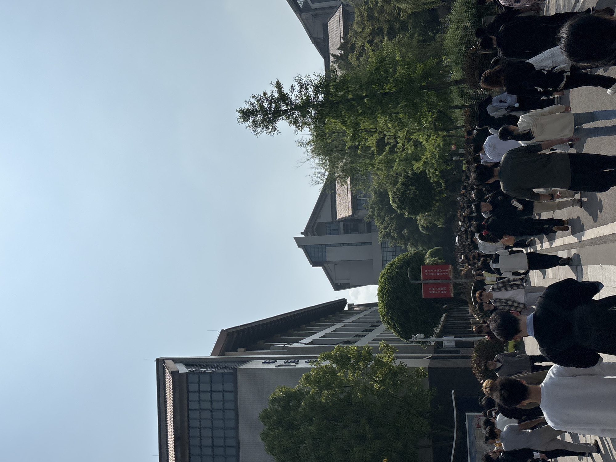 A large crowd of people is walking on a wide sidewalk next to a multi-story building in Jiangjin, China. The building has a distinctive roofline with a combination of traditional Chinese and modern architectural elements. Trees line the sidewalk, providing shade. The sky is blue and clear. A sign with Chinese characters is visible near the crowd. Most of the people appear to be young adults. They are walking in the same direction, suggesting a shared purpose or destination.