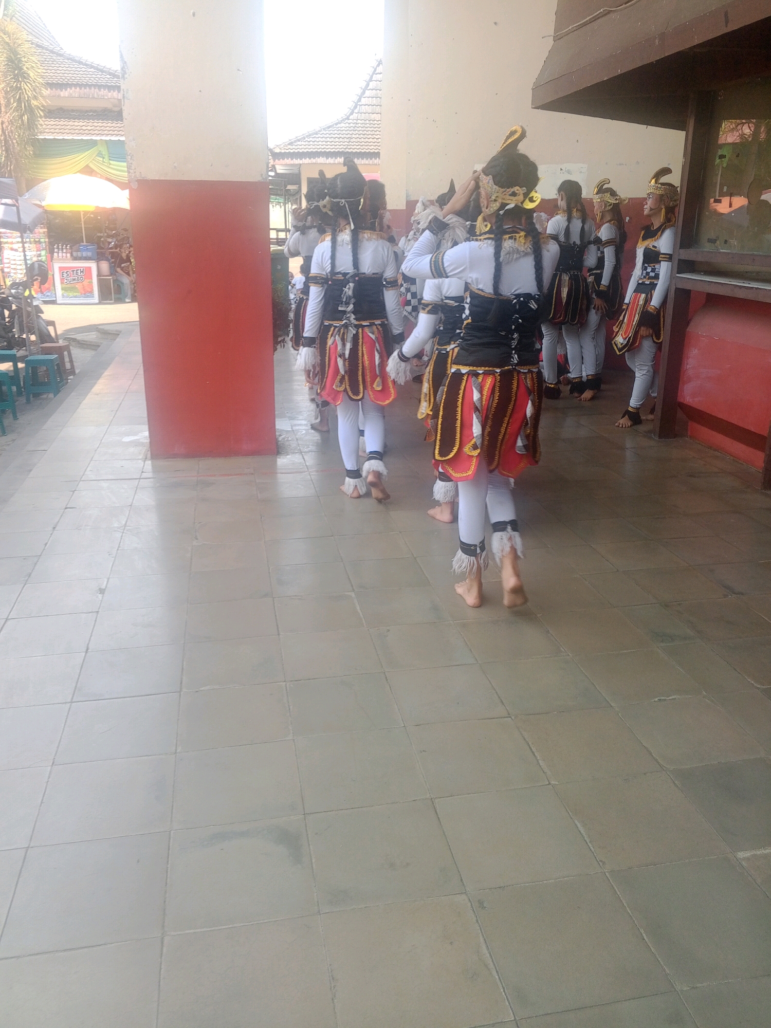 In Wuryorejo, Indonesia, a group of individuals in traditional attire are walking on a tiled pathway in front of a building. The individuals are wearing white tops, black vests, and layered red and gold skirts. Their hair is styled in long braids, adorned with golden headpieces. Some individuals have white fringe accents on their ankles. The pathway is made of light-colored square tiles. The background shows architectural elements of a building with red and white columns and a brown wooden structure. To the left, a market stall with a yellow and white awning is visible, displaying a sign that reads "ESTEH JUMBO." People are seated on green stools near the stall. The lighting suggests it is daytime, with sunlight casting shadows on the ground.