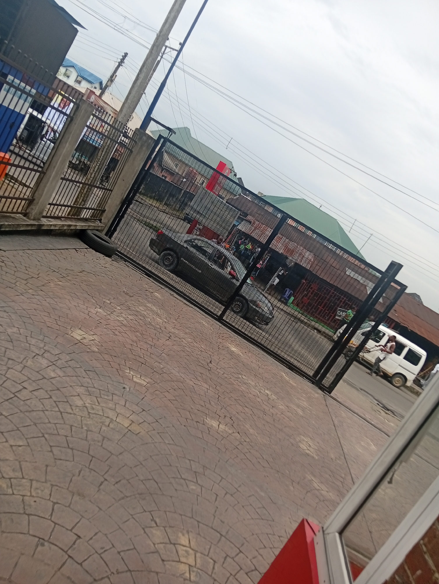 The image depicts a street scene in Rumuepirikom, Nigeria. In the foreground, a red structure, possibly a building or kiosk, is visible on the right side, reflecting in a glass surface. The ground is paved with reddish-brown interlocking bricks. A black metal fence runs diagonally across the scene, with a large tire placed against its base on the left.
Beyond the fence, several vehicles are present. A dark-colored sedan is parked behind the fence, and further down the street, a white minibus is visible, with two individuals near it. The background features buildings with varying roof colors, including green and reddish-brown, and utility poles with overhead wires. The sky is overcast and appears cloudy. The overall impression is a moderately busy urban environment.