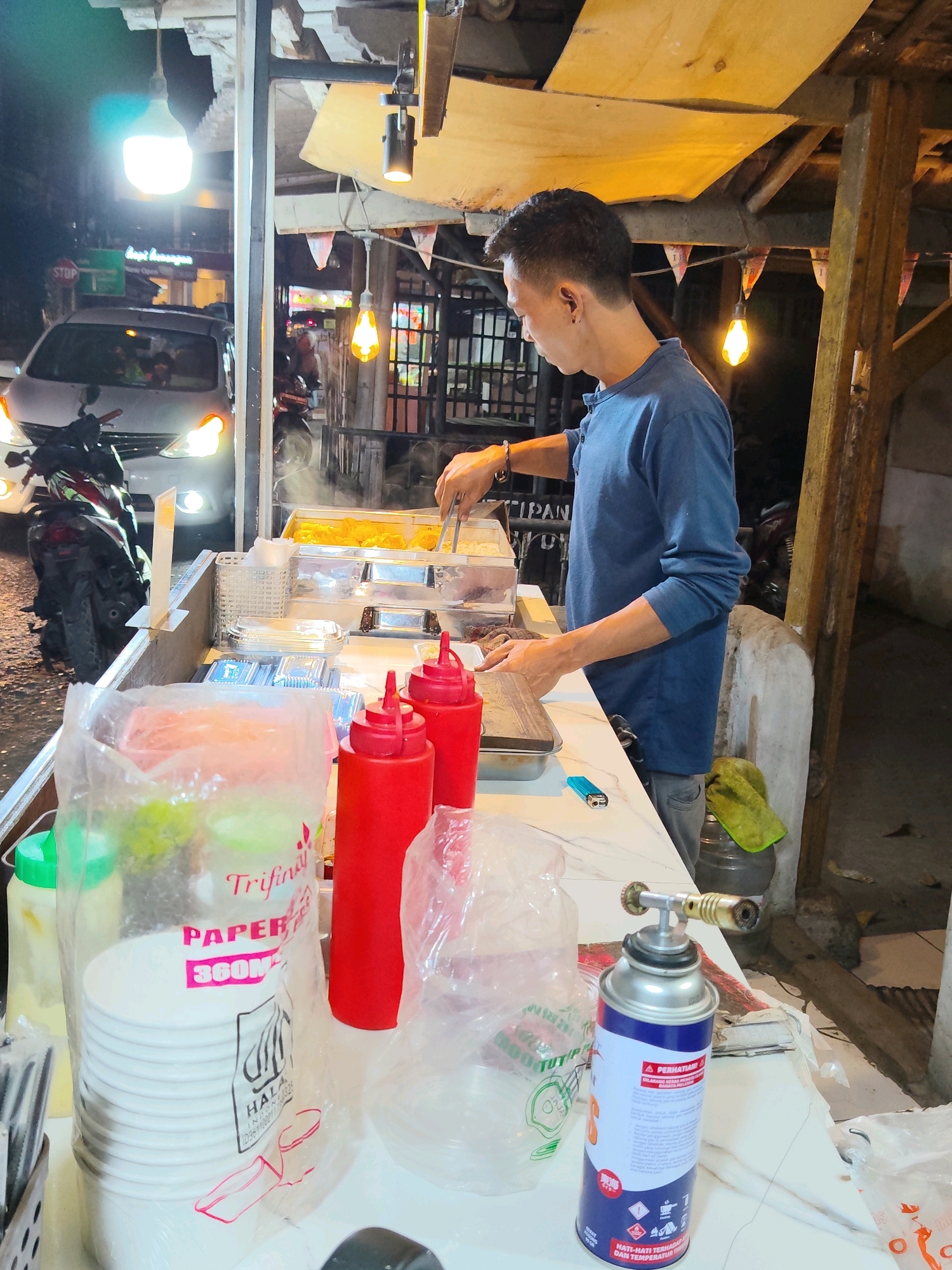 A young man in a blue long-sleeved shirt is preparing food at a street food stall in Purwakarta, Indonesia. The stall is set up outdoors at night, with overhead lights illuminating the scene. In the background, vehicles and other stalls are visible. The man is using tongs to serve food from a metal container, while other food containers and condiments are arranged on the counter. There are stacks of paper bowls, red squeeze bottles, and a butane torch on the table. The overall impression is of a busy night market or street food operation.