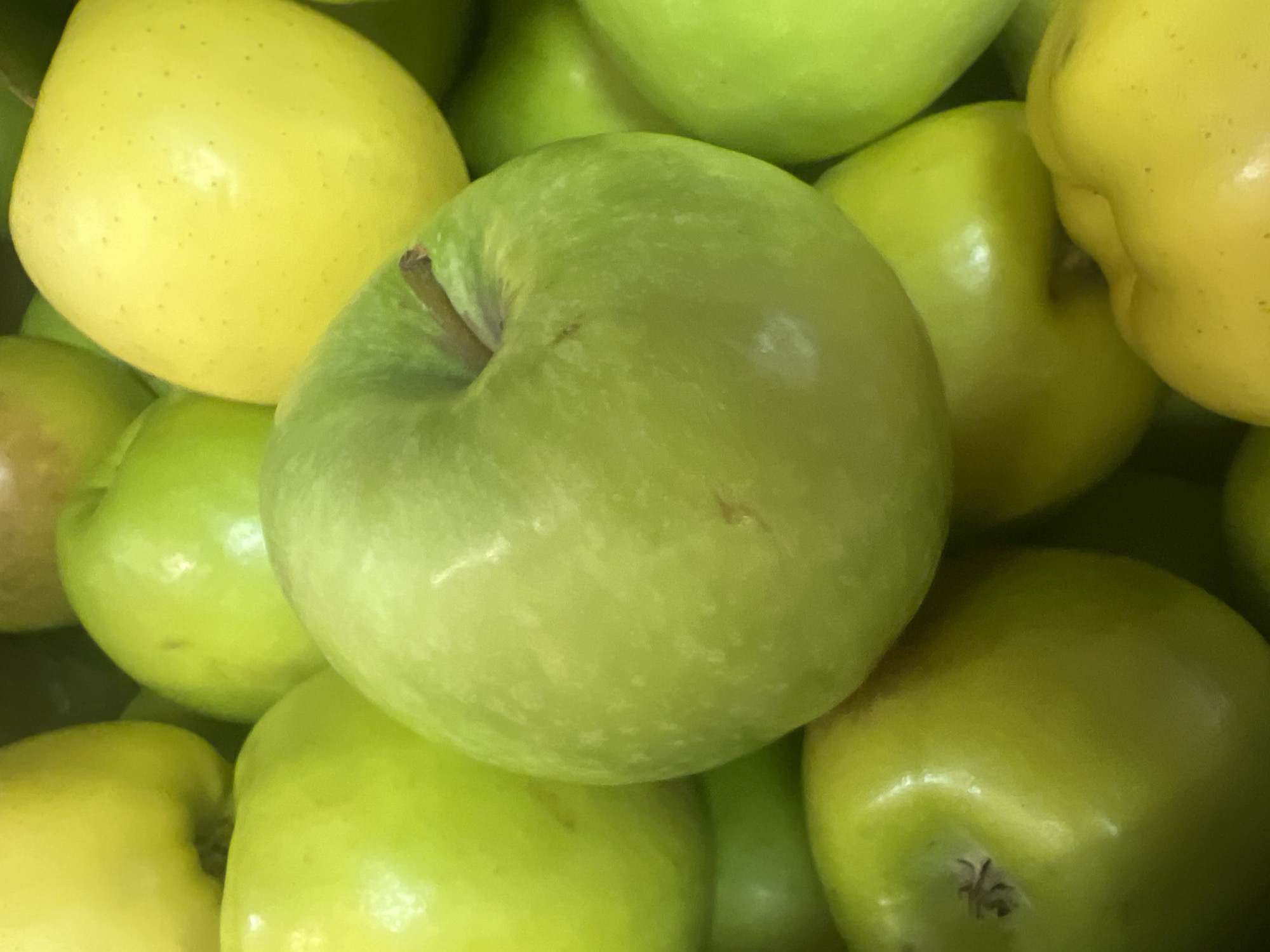 A close-up, eye-level, indoor shot shows a pile of ripe, green apples with a few yellow apples interspersed. The apples are arranged tightly together, filling the frame. The lighting is bright and natural, likely from a storefront or market. The image is devoid of human presence, traffic, or any recognizable landmarks. The absence of specific urban identifiers makes it impossible to pinpoint a location within Johannesburg, South Africa.