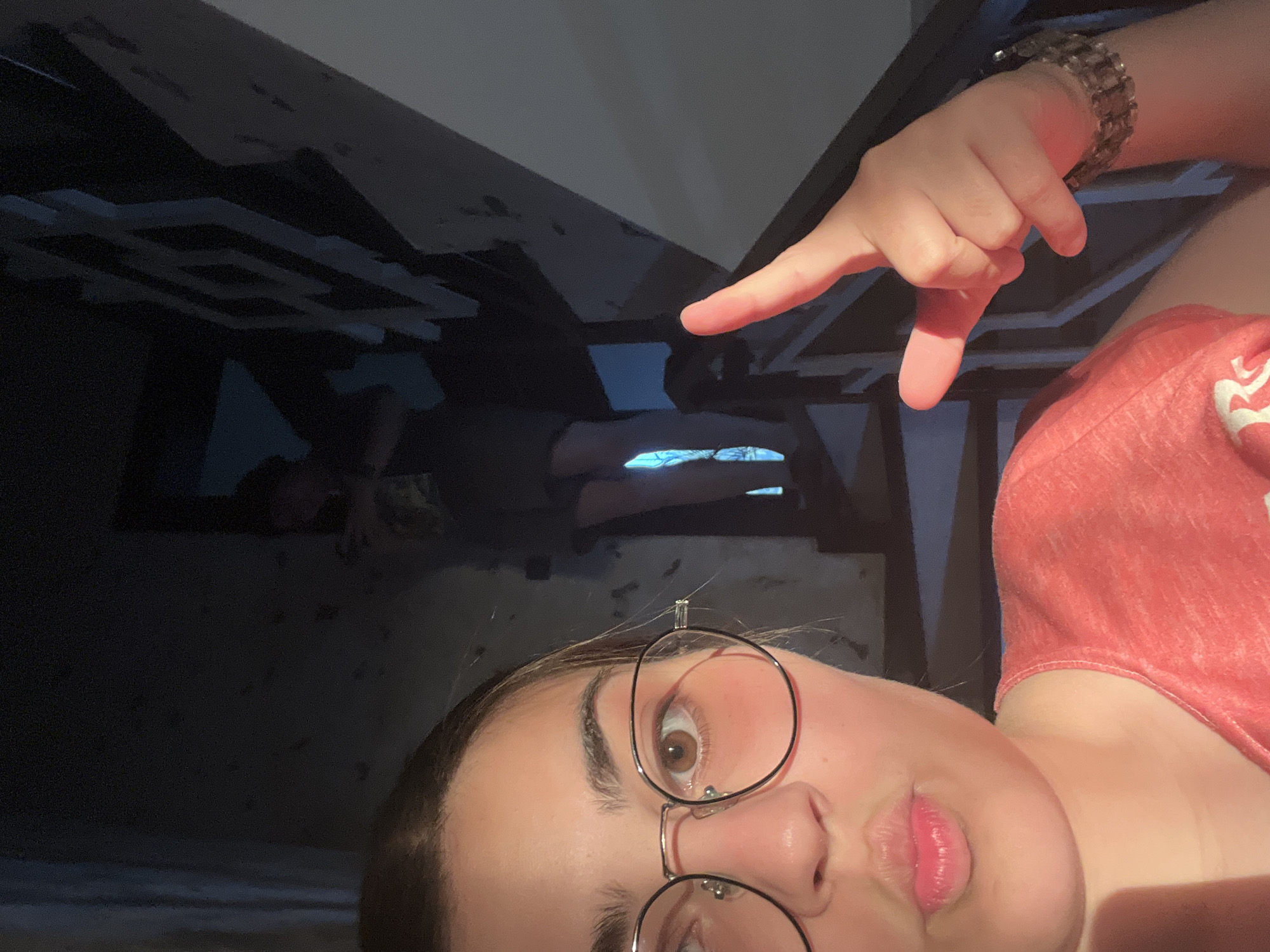 The image shows a young woman taking a selfie in a dimly lit room with a reflective floor. She is wearing a pink t-shirt and glasses. Her left hand is raised, with her index finger extended, pointing towards the reflective surface. In the reflection, another person can be seen lying down. The room appears to be indoors, with geometric patterns visible on the floor and walls, possibly indicating a maze-like structure or patterned tiling. The lighting is low, with some light sources creating reflections on the floor. The overall mood is casual and playful, with the woman's expression suggesting a lighthearted moment. There is no visible text in the image. The location context is Koroviia, Ukraine.