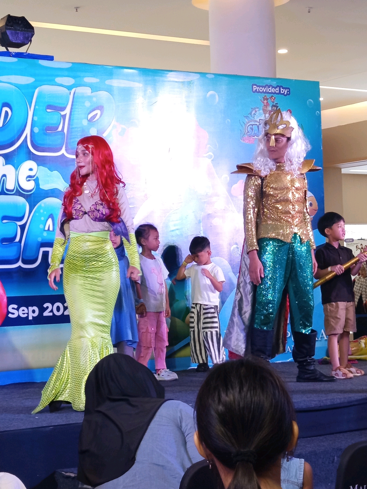 This image captures a performance on a stage in Ciakar, Indonesia, likely at an indoor venue like a mall, given the lighting and suspended lights. A woman dressed as a mermaid with a red wig and a green scale-patterned skirt is standing on the left. To her right, a person in a costume resembling King Triton, with a gold sequined top, teal sequined pants, and a white wig, stands tall. Several children are also present, some watching the performance and others appearing to be part of it. One boy on the far right is holding a golden prop. The backdrop features a blue design with "DEEP THE SEA" and "Provided by:" text, suggesting an event themed around the ocean. The overall atmosphere appears festive, with people dressed in elaborate costumes for a public event.
