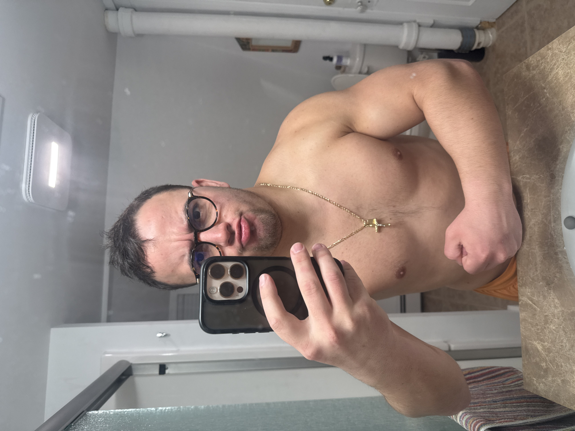 A shirtless man with dark hair and glasses is taking a selfie in a bathroom. He is wearing a gold chain with a cross pendant. His right arm is flexed. The bathroom has white walls and a marbled countertop. A towel and a sink are visible. A light fixture is on the wall. The location is likely not in Provincetown, United States of America, as there is no information to support that claim.