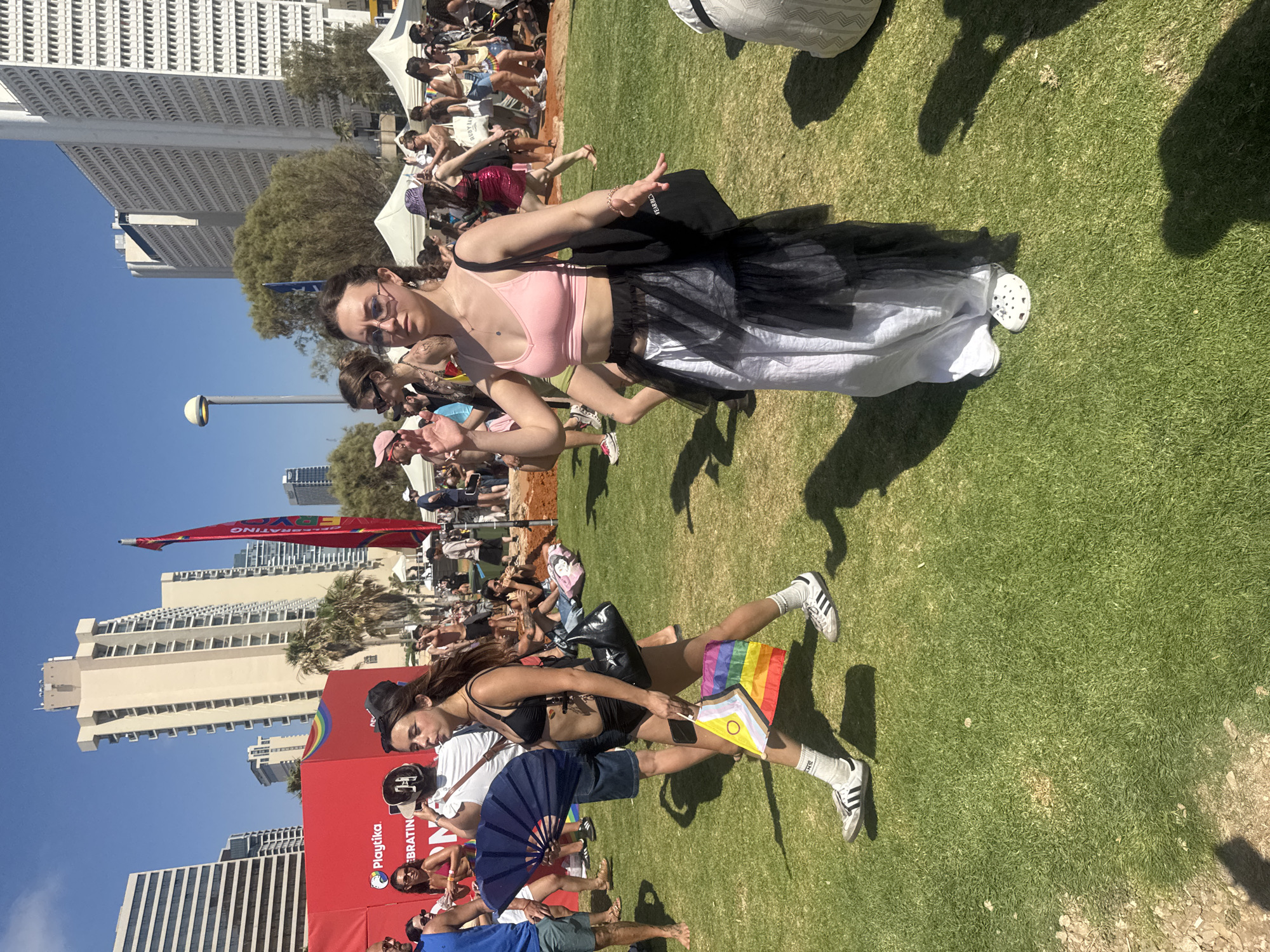 This media file shows a crowd of people at an outdoor event, likely a festival or parade, on a sunny day. The scene is set on a well-maintained grassy area with several modern buildings visible in the background, suggesting an urban location. The architecture of the buildings, along with palm trees, hints at a warm climate.
The subjects are primarily young adults, with a focus on several women in the foreground and middle ground. One woman in the foreground is wearing a black bikini top, denim shorts, and white sneakers, and is holding a small rainbow flag. Another woman in the middle ground is dressed in a pink crop top and a black and white skirt, with her arms extended. People in the background appear to be watching or participating in the event, some sitting on the grass and others standing.
A prominent red banner with white lettering is visible, displaying text that includes "Playtika" and "CELEBRATING." A rainbow graphic is also incorporated into the red banner. Another flag, a rainbow flag, is held by a person in the foreground. The atmosphere appears festive and celebratory, with people smiling and engaging in the event. The time of day is daytime, indicated by the bright sunlight and clear blue sky.