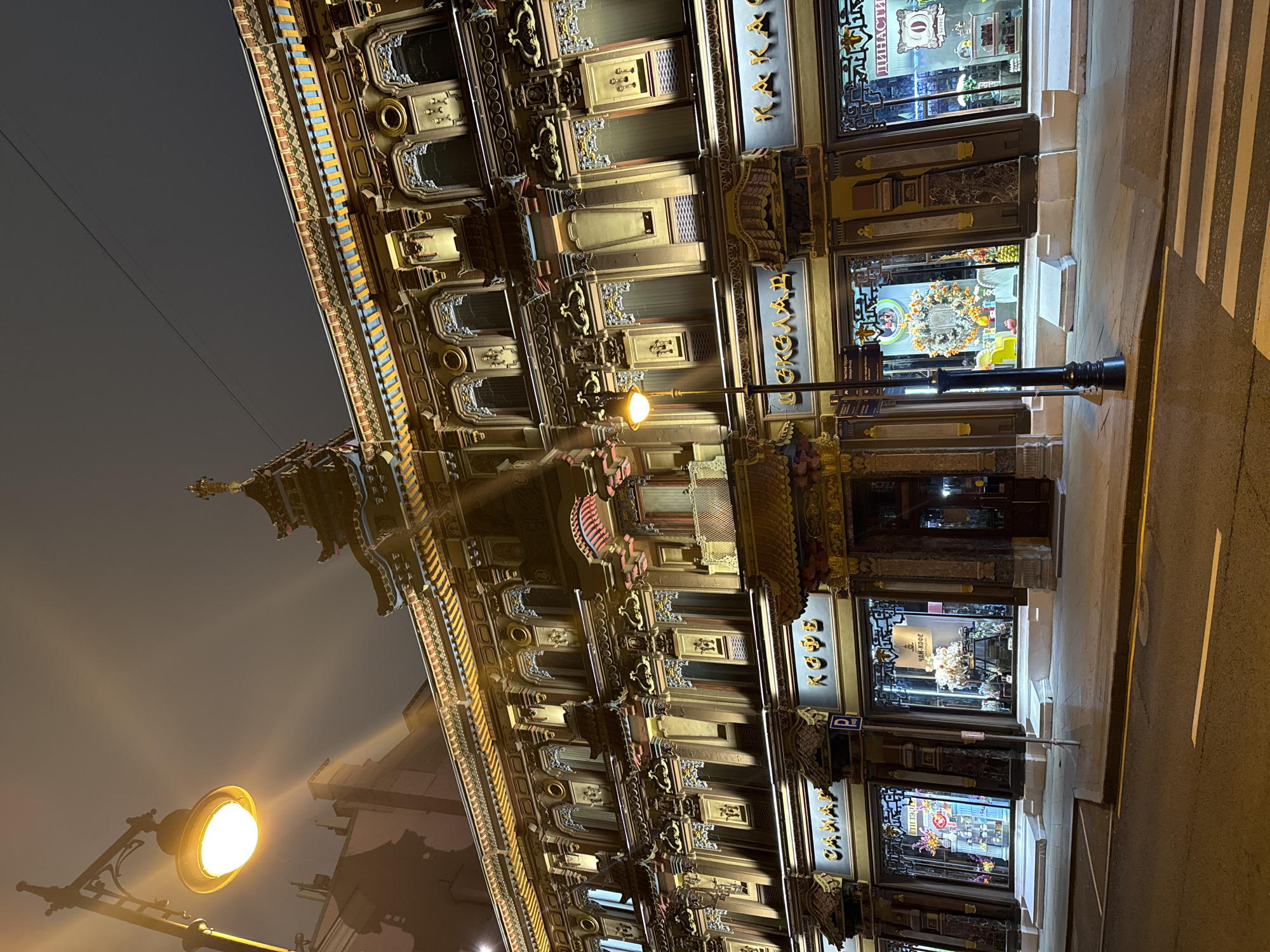 The image depicts a building in Moscow, Russia, illuminated at night. The architecture is ornate, with multiple levels and decorative elements. The building's facade is adorned with carvings, patterns, and trim. Shopfront windows line the ground level. A lamppost stands near the building, providing light. The street is visible, marked with a pedestrian crossing. The sky is dark, suggesting it is evening or night.